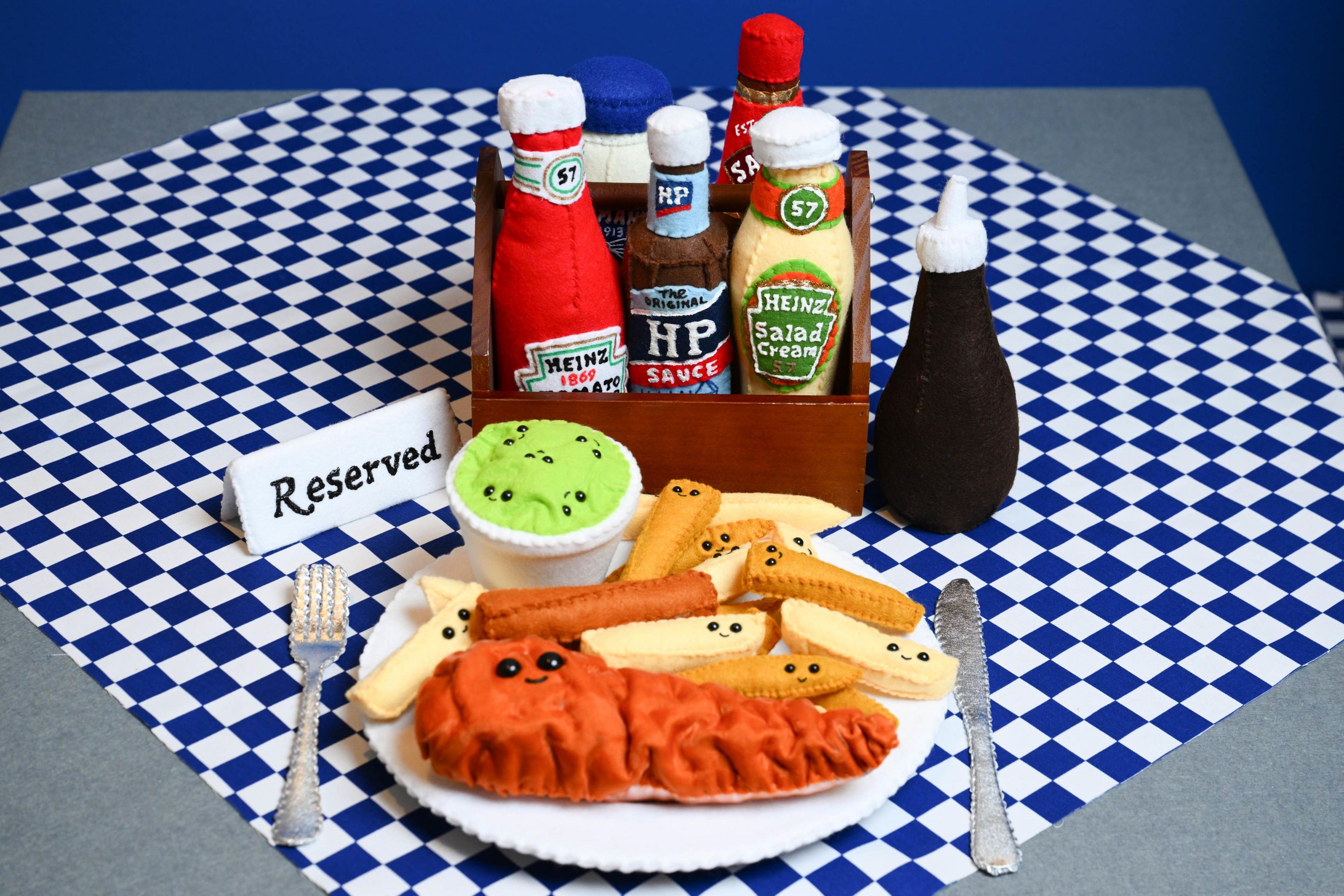 A plate of fish, chips and mushy peas with smiley faces on them, with condiments including ketchup, brow sauce and salad cream behind it.