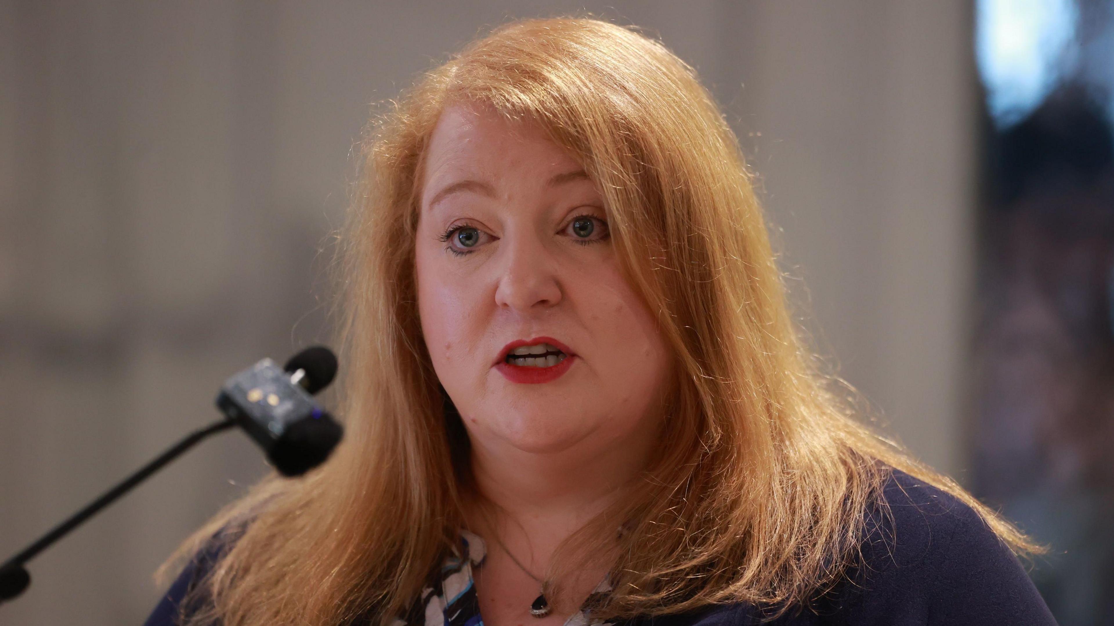 Naomi Long with shoulder length ginger hair and red lipstick as she wears a navy, white, blue and purple top.