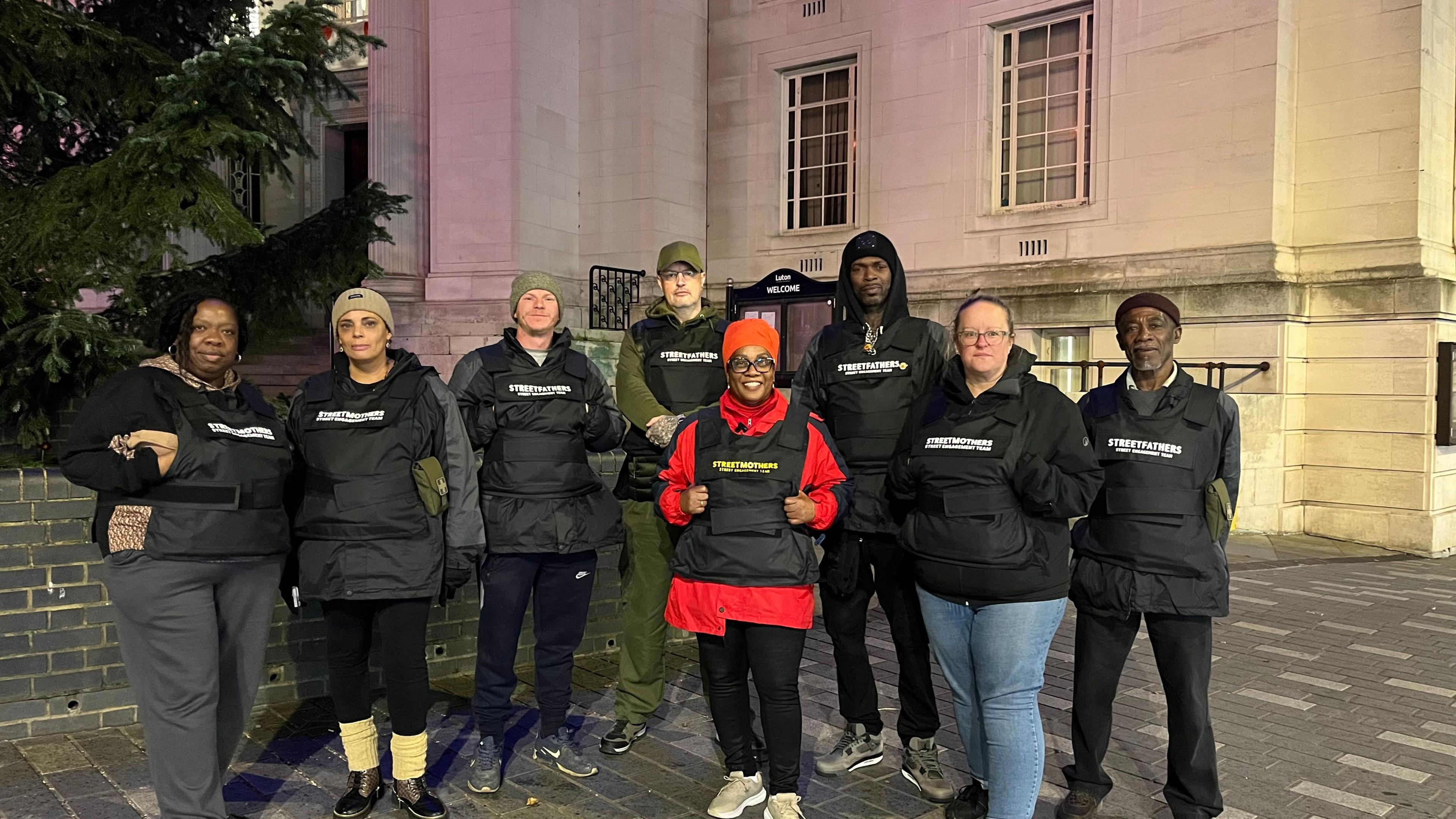 A group of eight volunteers are standing together outdoors in front of a large stone building with tall windows and steps leading up to an entrance. The volunteers are wearing black jackets and black stab-proof vests with white text that reads “Street Fathers” or “Street Mothers.” One person in the centre stands out in a bright red coat under the vest.