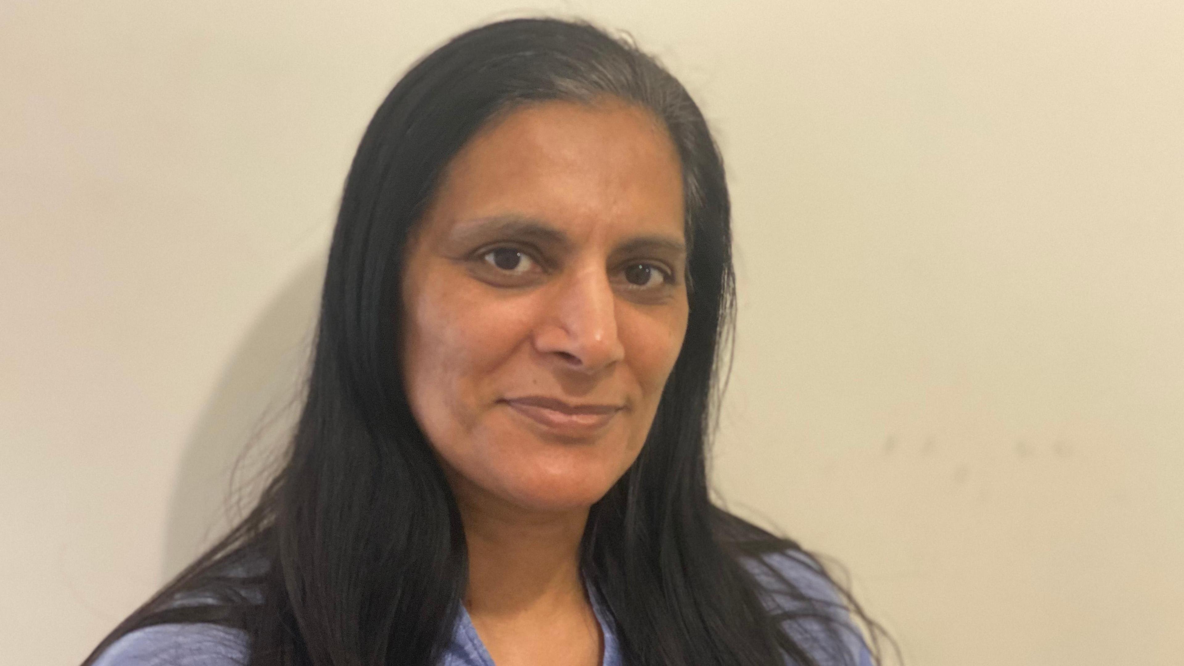 Rashida Anwar with long black hair wearing a blue top with a neutral expression standing against a plain cream wall