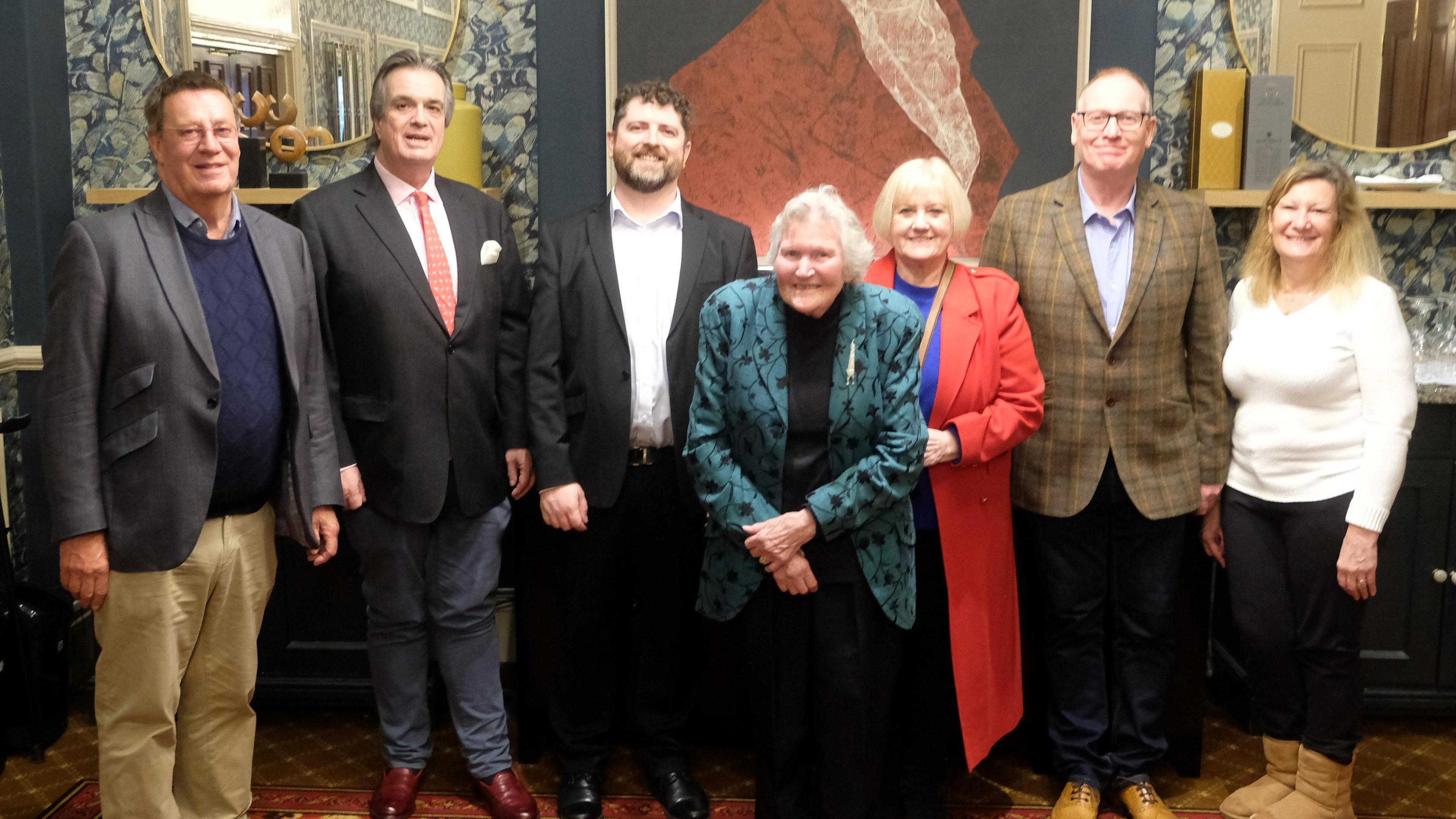 A group of seven people in a room, including Gill Green. There are four men, including the Duke of Bedford. The men are all in smart clothes, including jackets. A woman in a red coat is standing behind Mrs Green. Another woman to the right is wearing a white top, black trousers and beige boots. They are all smiling. 