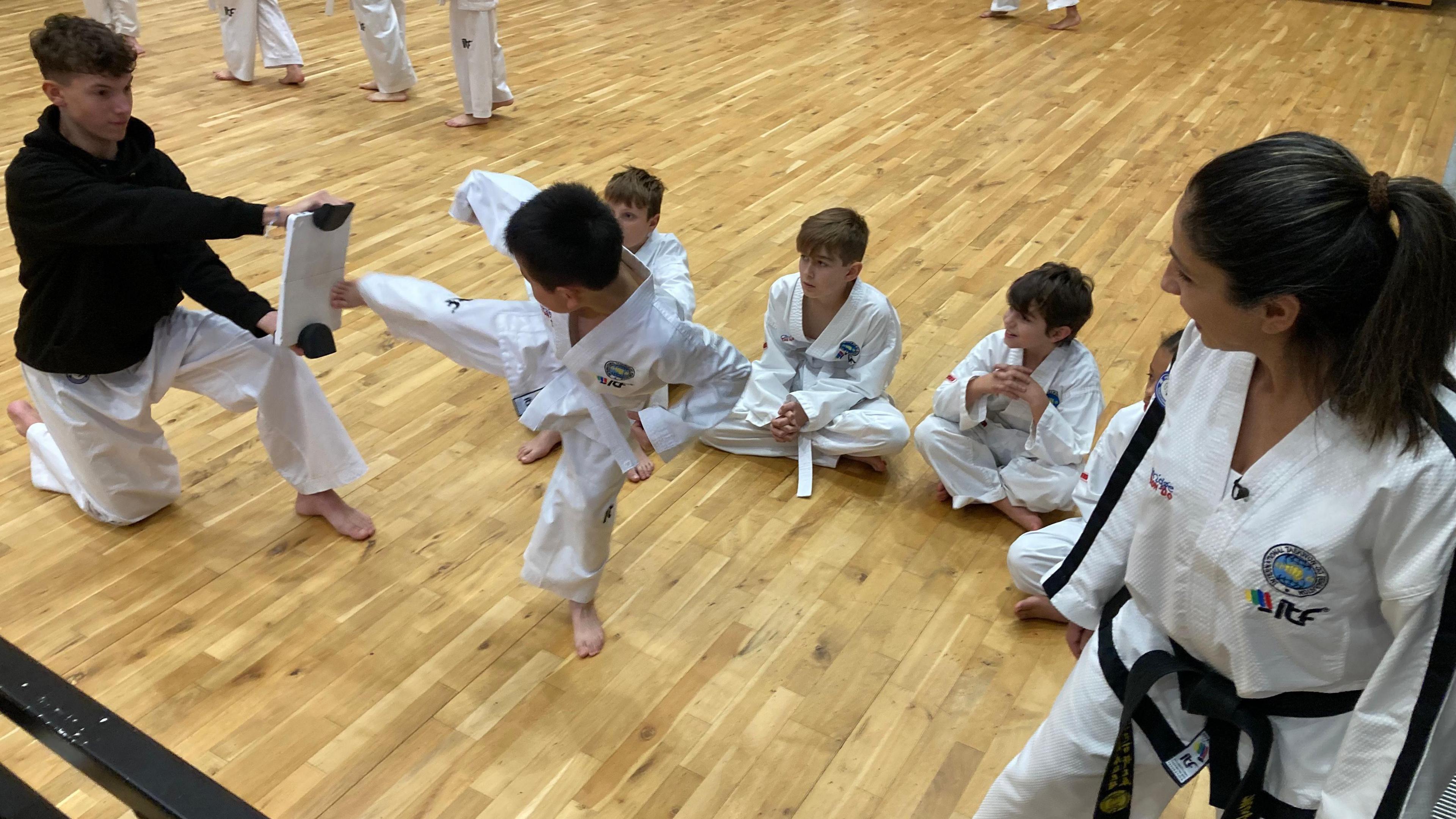 One of the club's younger members demonstrates his taekwondo kick on a pad being held by a teenager. Other youngsters, also wearing martials arts suits, sit cross-legged on the floor. A female adult instructor wearing a black belt looks on.