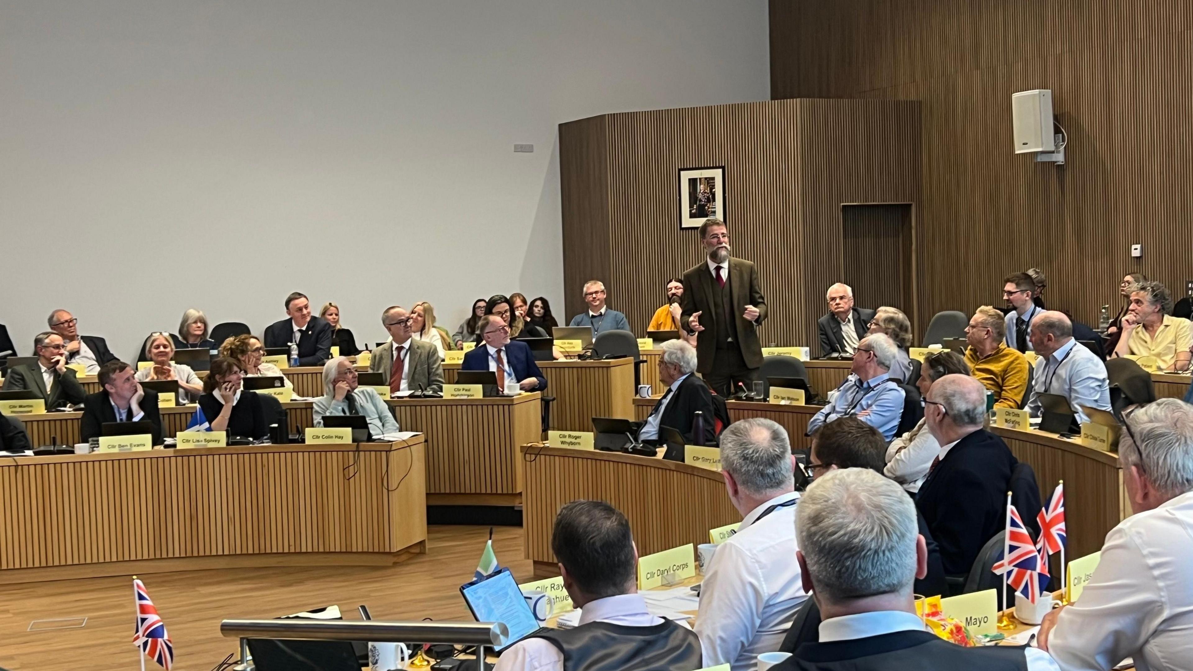 A large group of councillors sit at desks in a circular council hall. Some have union jack flags on their desks. One man in a tweed suit with grey hair and a beard is standing up speaking. Everyone is looking at him. Many councillors are only visible from the back. 