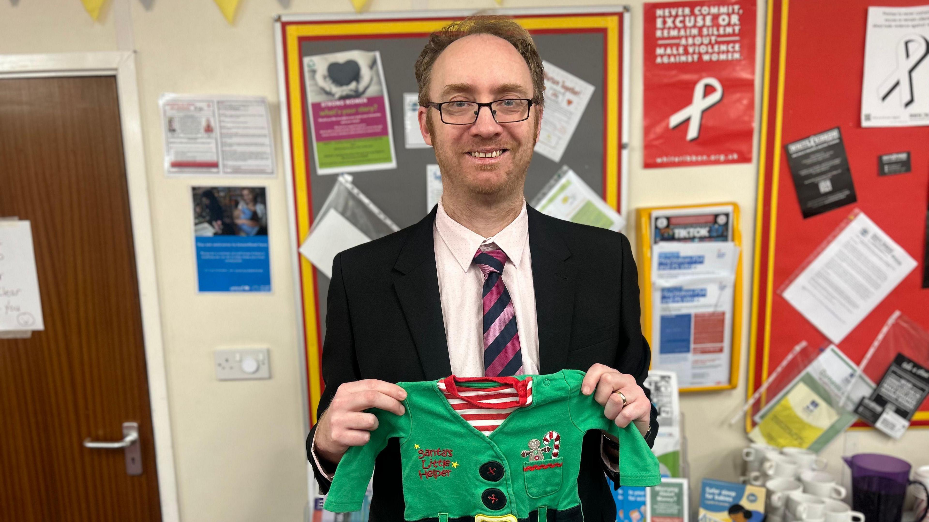 Councillor Charles Quinn stands in a school corridor and is holding a small green, red and white Christmas jumper in the style of an elf outfit. He is wearing a dark suit, white shirt and a pink, purple and blue striped tie. He has a stubble beard and is wearing glasses.