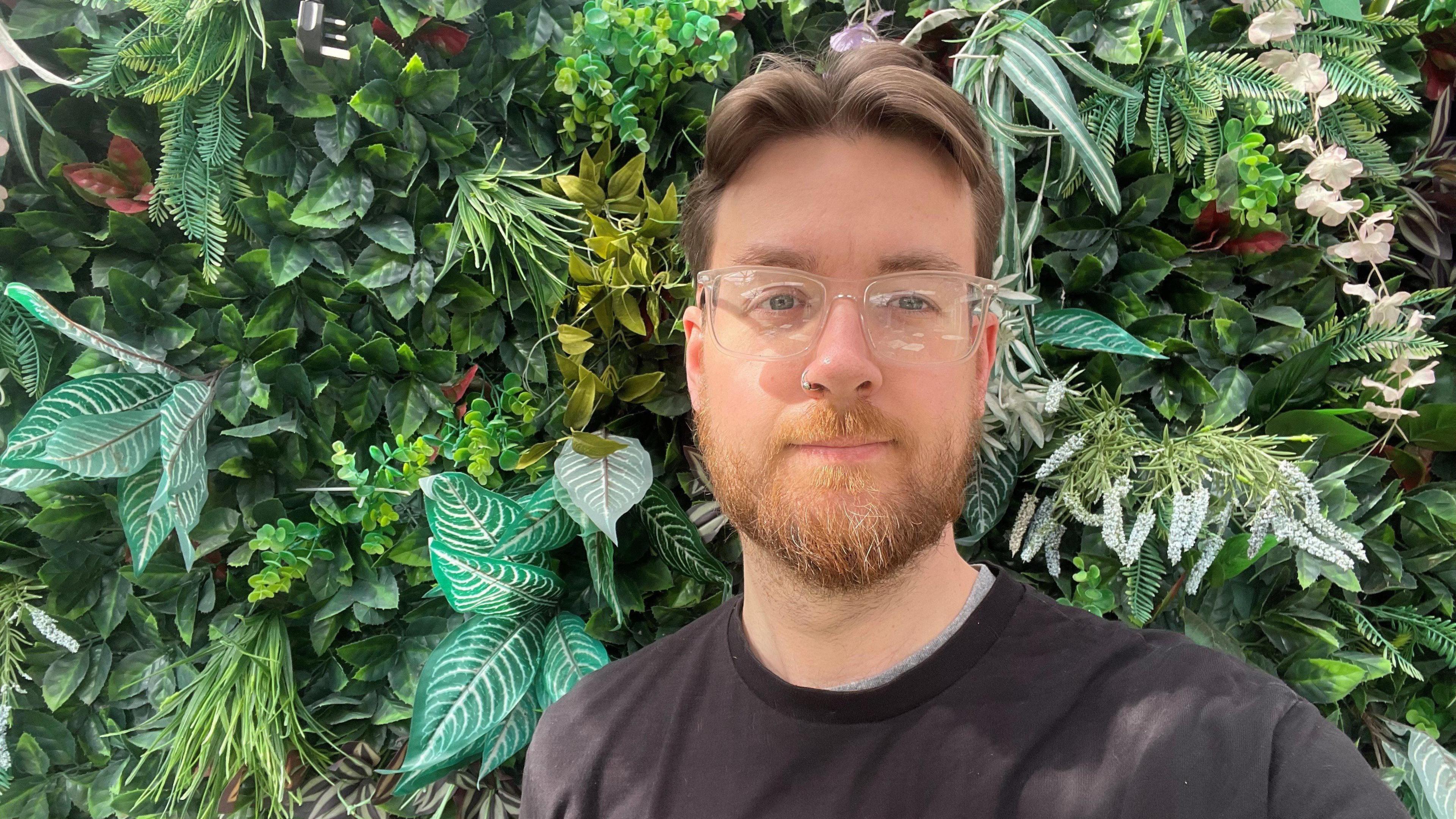 A man with short brown hair and beard is wearing clear framed glasses and a black T-shirt. He is standing in front of greenery.