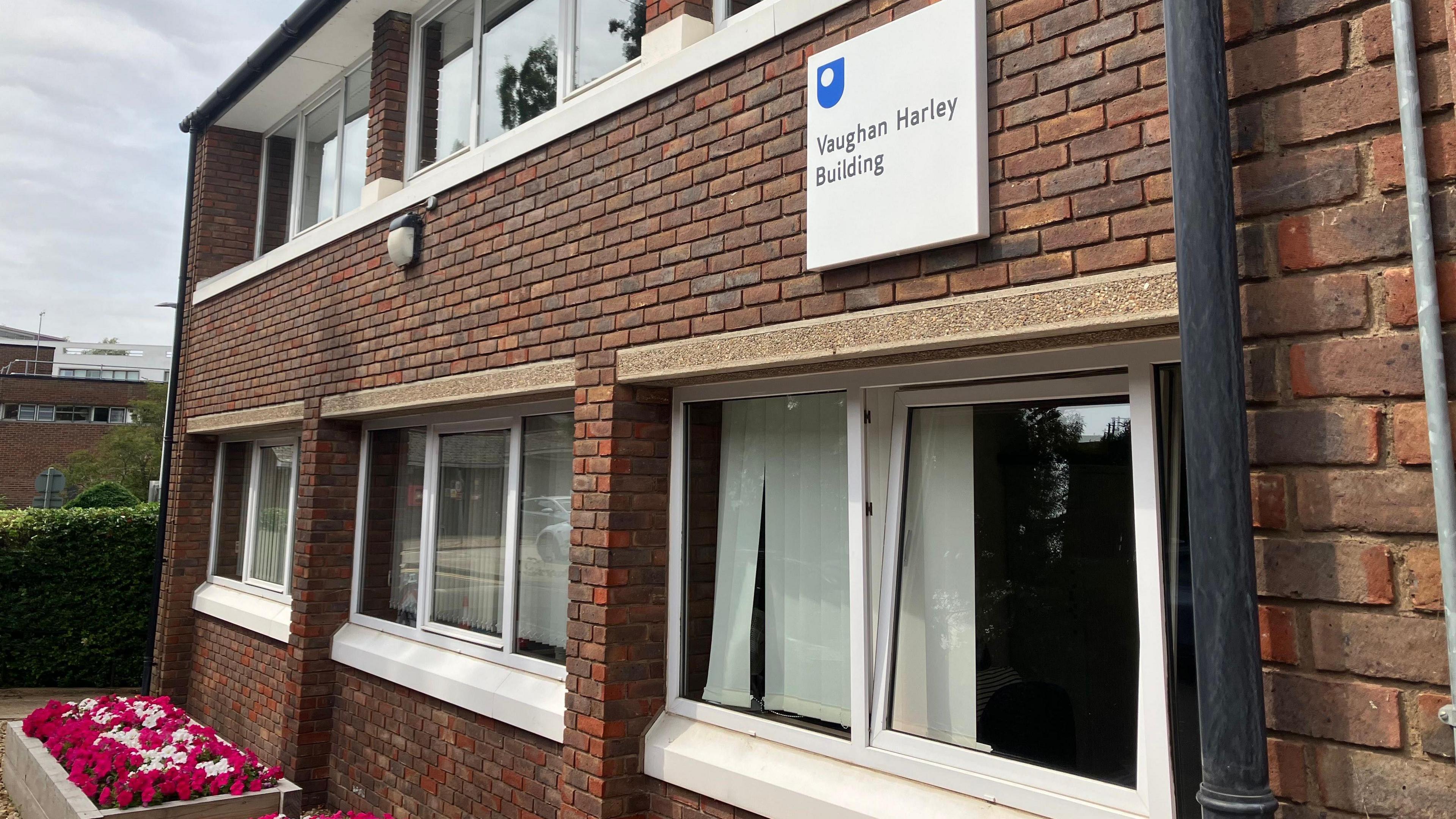 A picture of the Vaughan Harley building at the Open University in Milton Keynes, which is the current base for Healthwatch Milton Keynes. It is a two storey building with windows and you can see a flowerbed with red and white flowers in front of it.