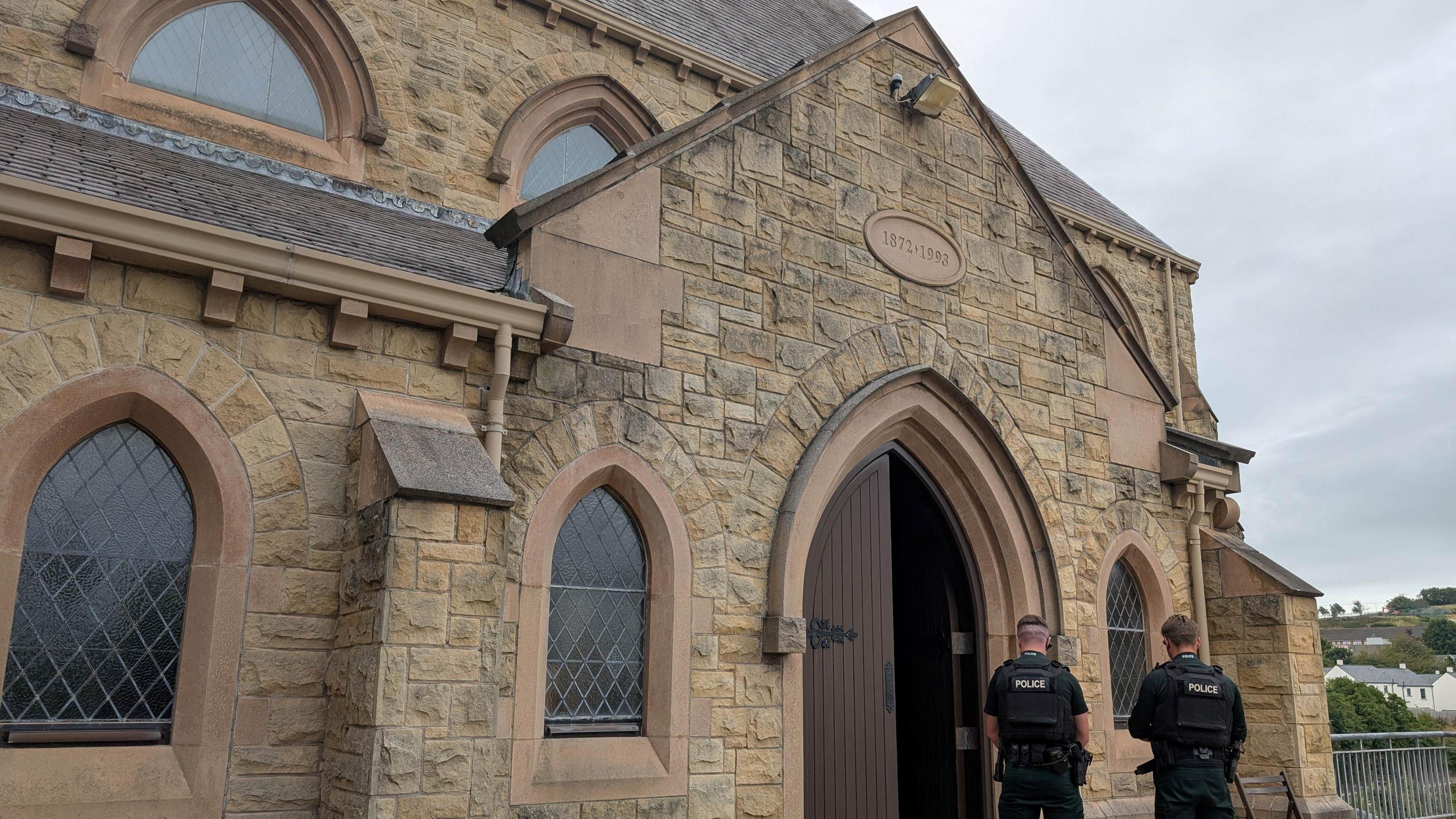 St Patrick’s Church in Downpatrick. It is an old brown building with a large brown door. Two PSNI officers are stood in front of the door with the backs to the camera. The sky is overcast.