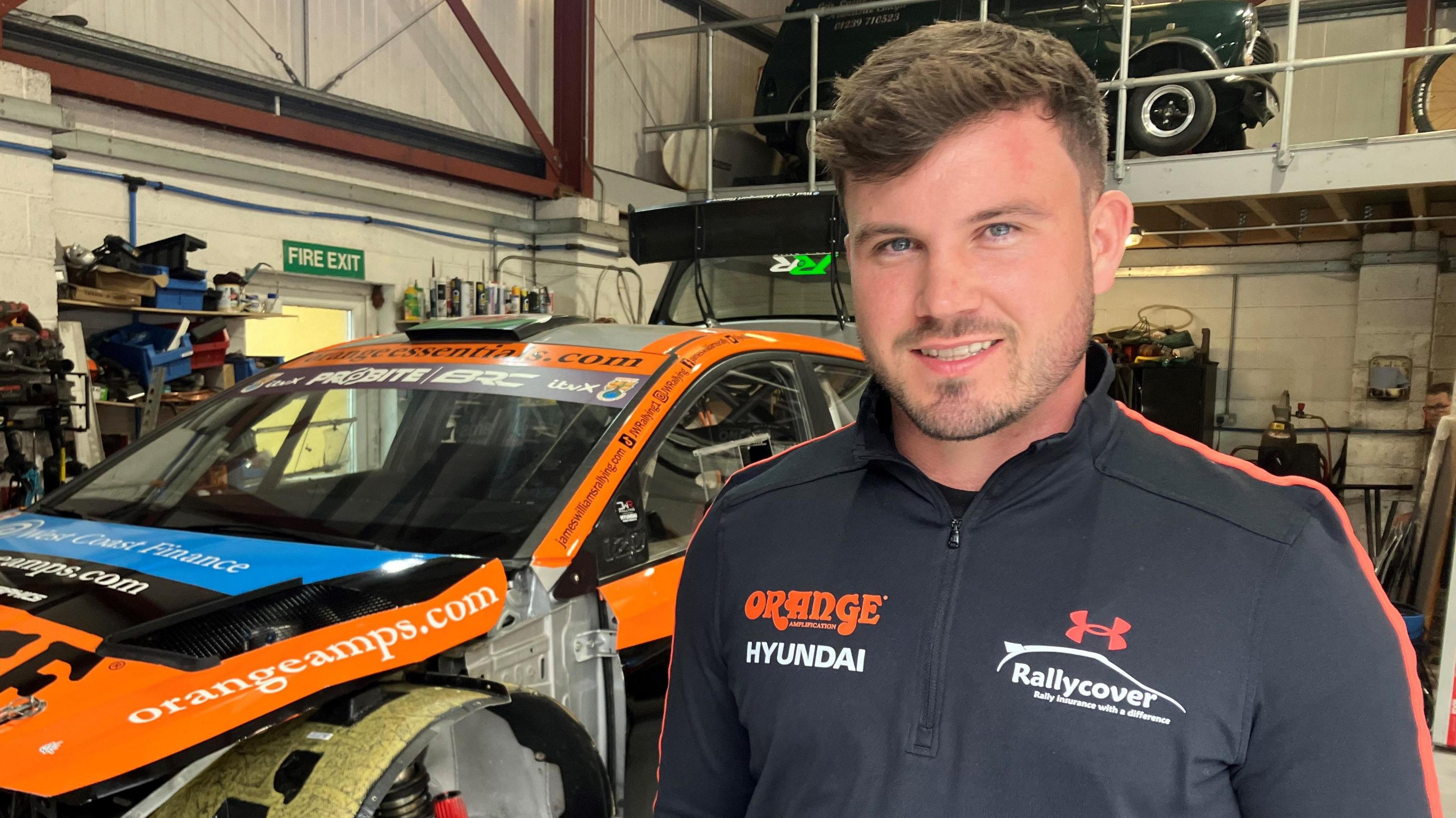 A young man with light brown hair, head and shoulders shot, standing in front of an orange rally car in a garage full of tool.