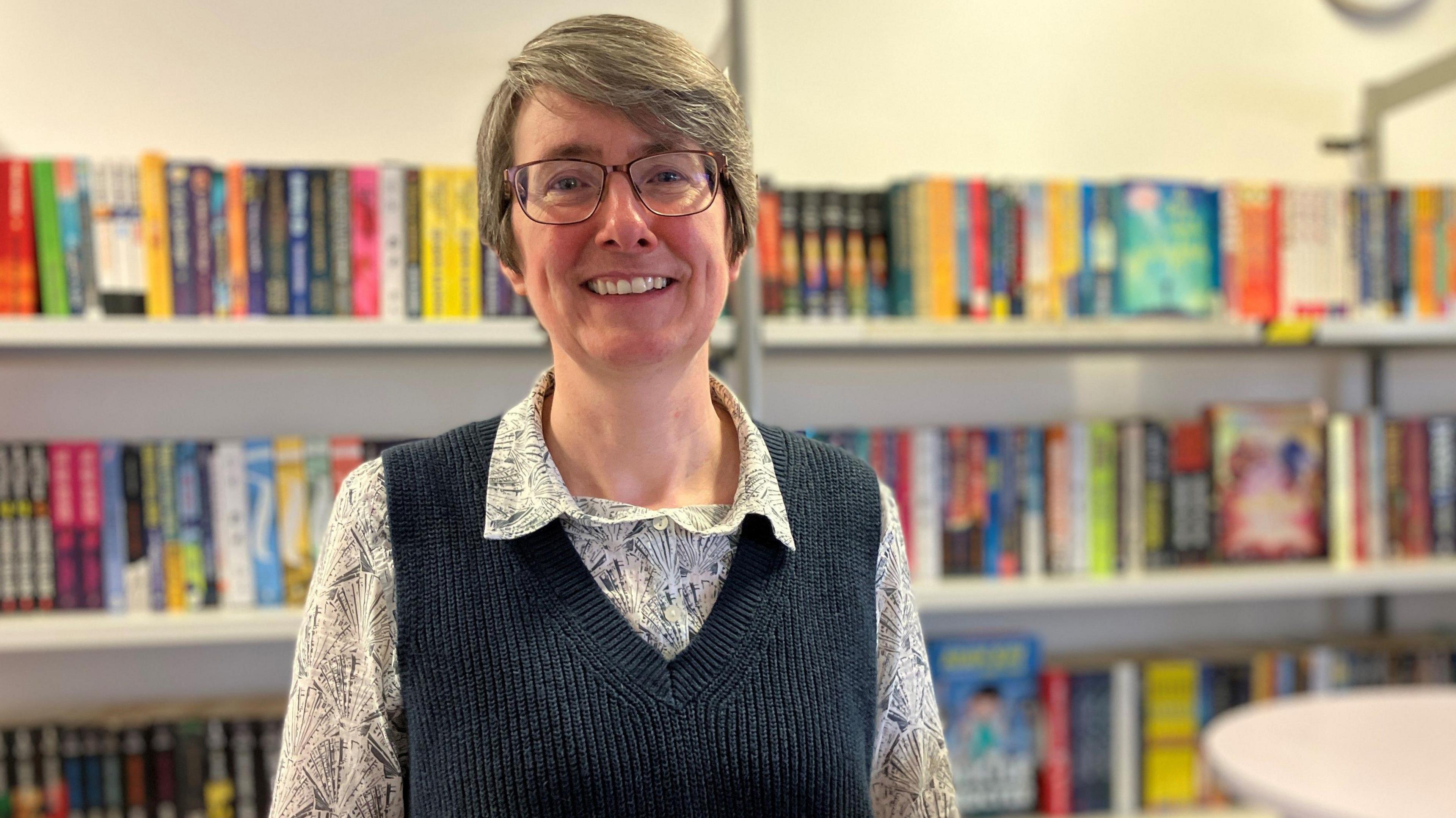 Durham Johnston School's Latin teacher Cathy Bothwell wears a navy v-neck vest and a patterned shirt. She stands in front of hundreds of book stacked on shelves. 