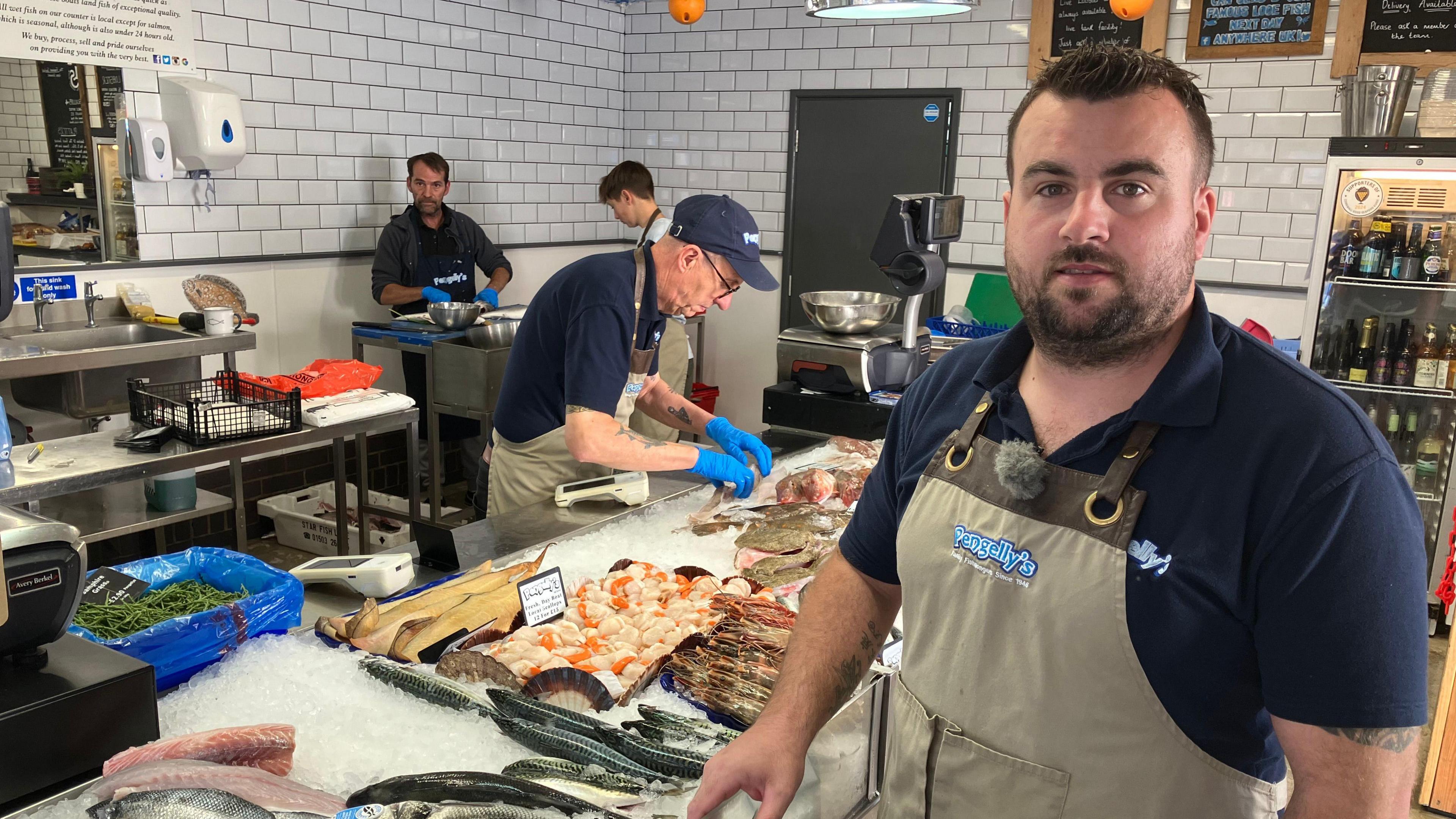 A man wearing an apron in a fishmongers.  Behind the man is a table covered in fish on ice and workers in aprons, preparing fish. 