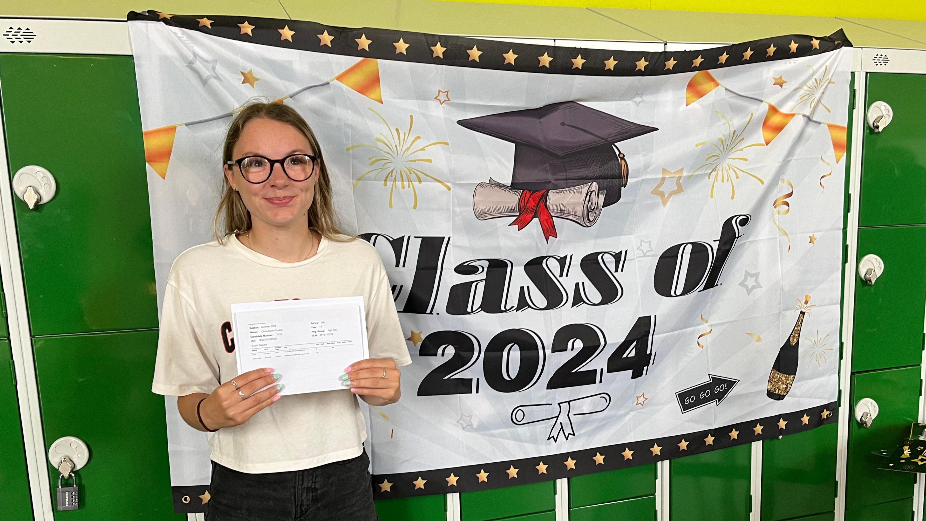 Olivia stands in front of a banner saying "class of 2024" holding her results envelope. She is wearing black glasses, has long blonde hair and wears a white t-shirt and black shorts. 