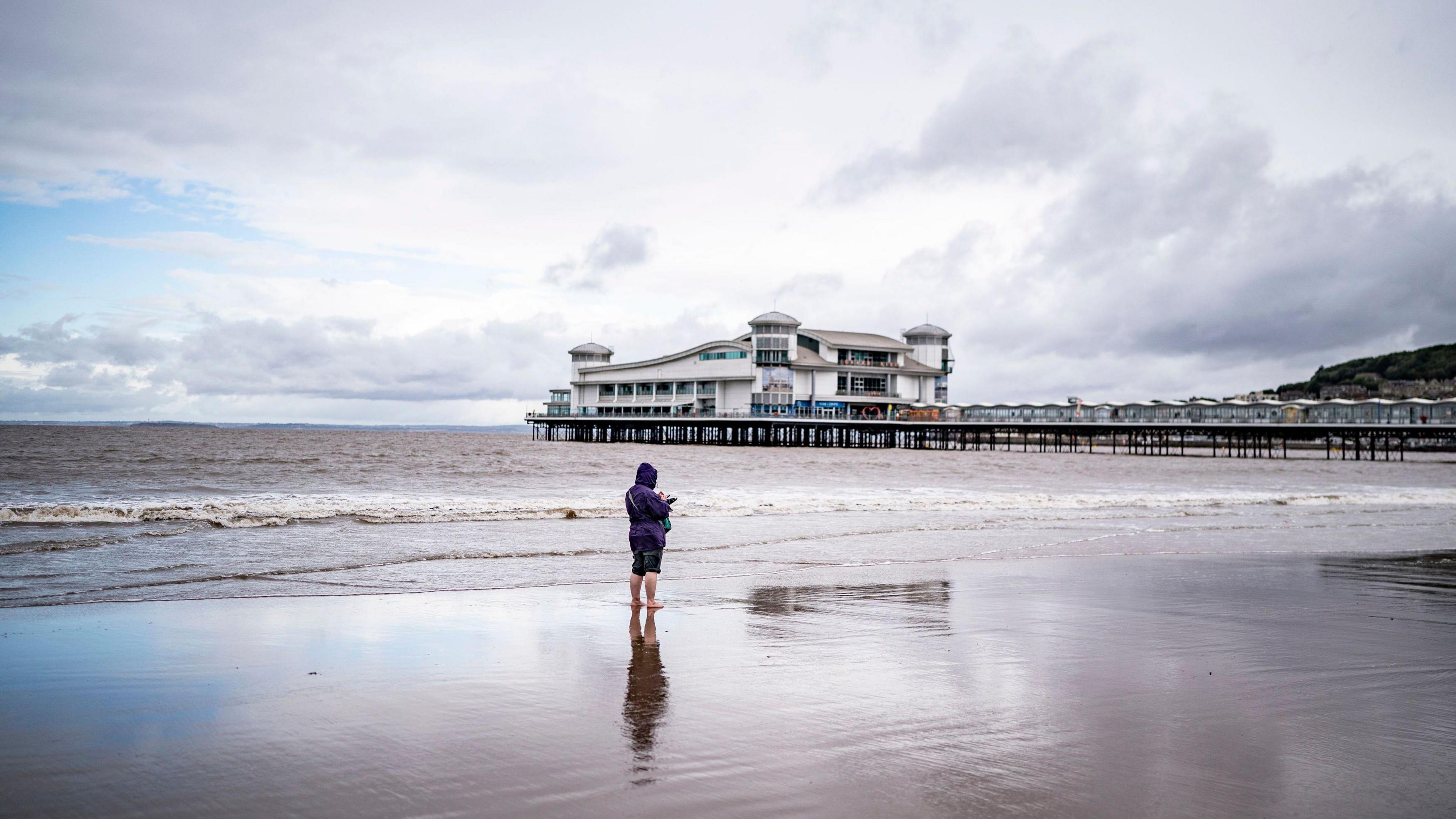 A person wearing a water proof with the hood up is standing by the sea in front of Weston-super-Mare's pier. The sky is grey, reflecting grey in the water.