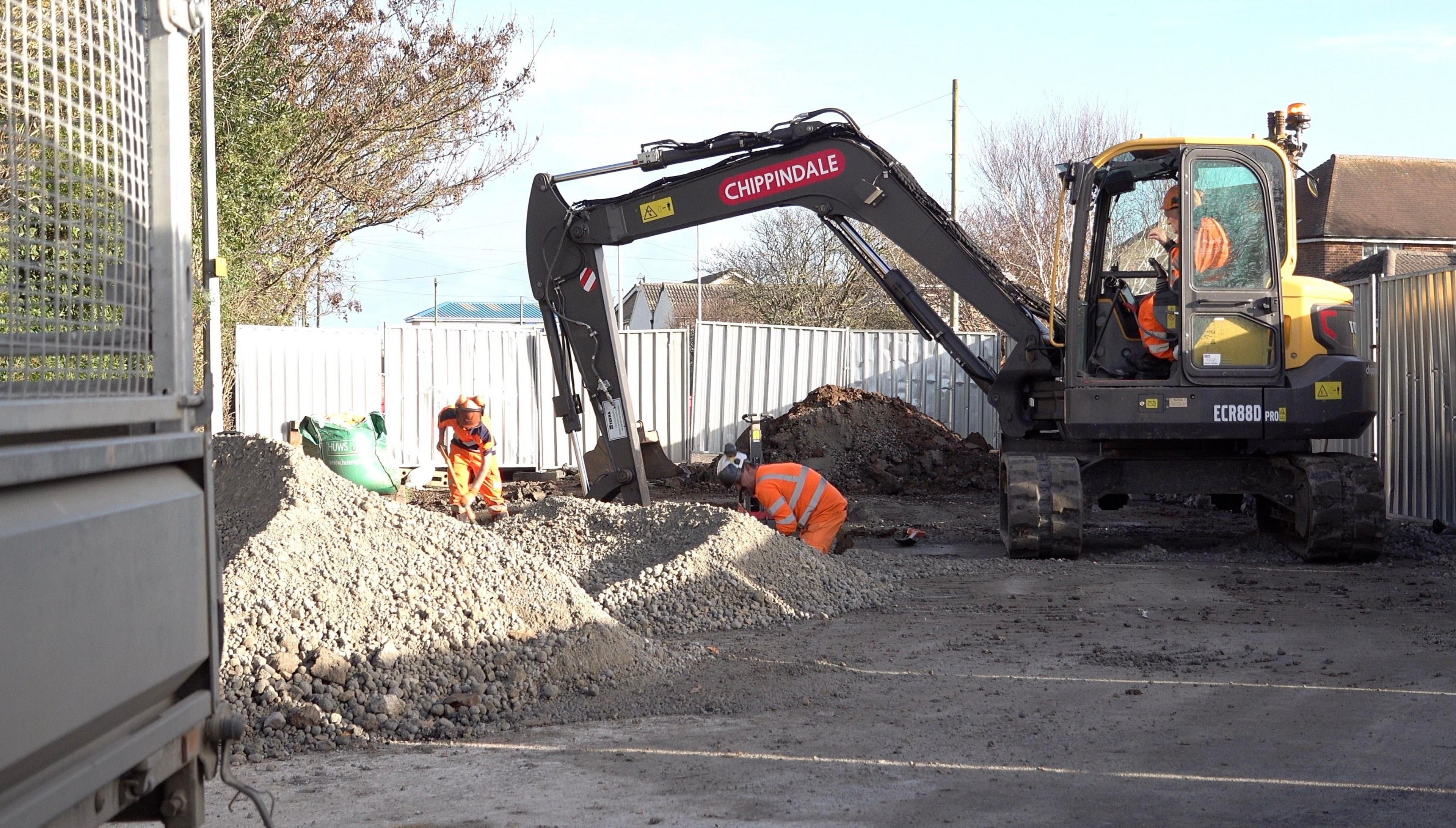 Three workers in orange, high-vis clothing carry out repairs to a road surrounded by rubble. One of the workers is inside a large digging machine while the two others are using hand tools.