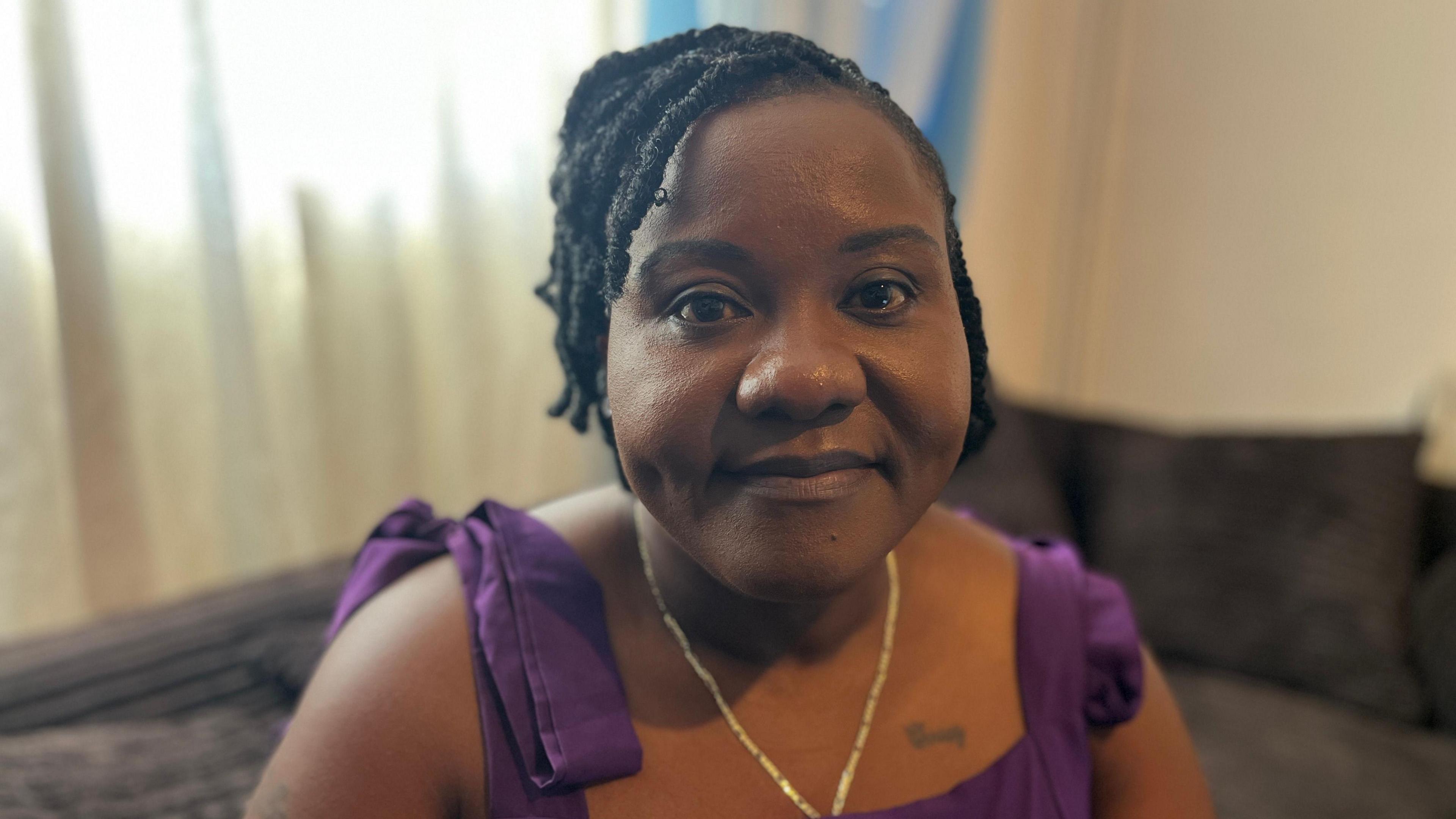 Christiana Hansen is looking straight into the camera, she is sitting on a sofa in her living room. She is wearing a purple dress and a gold chain.