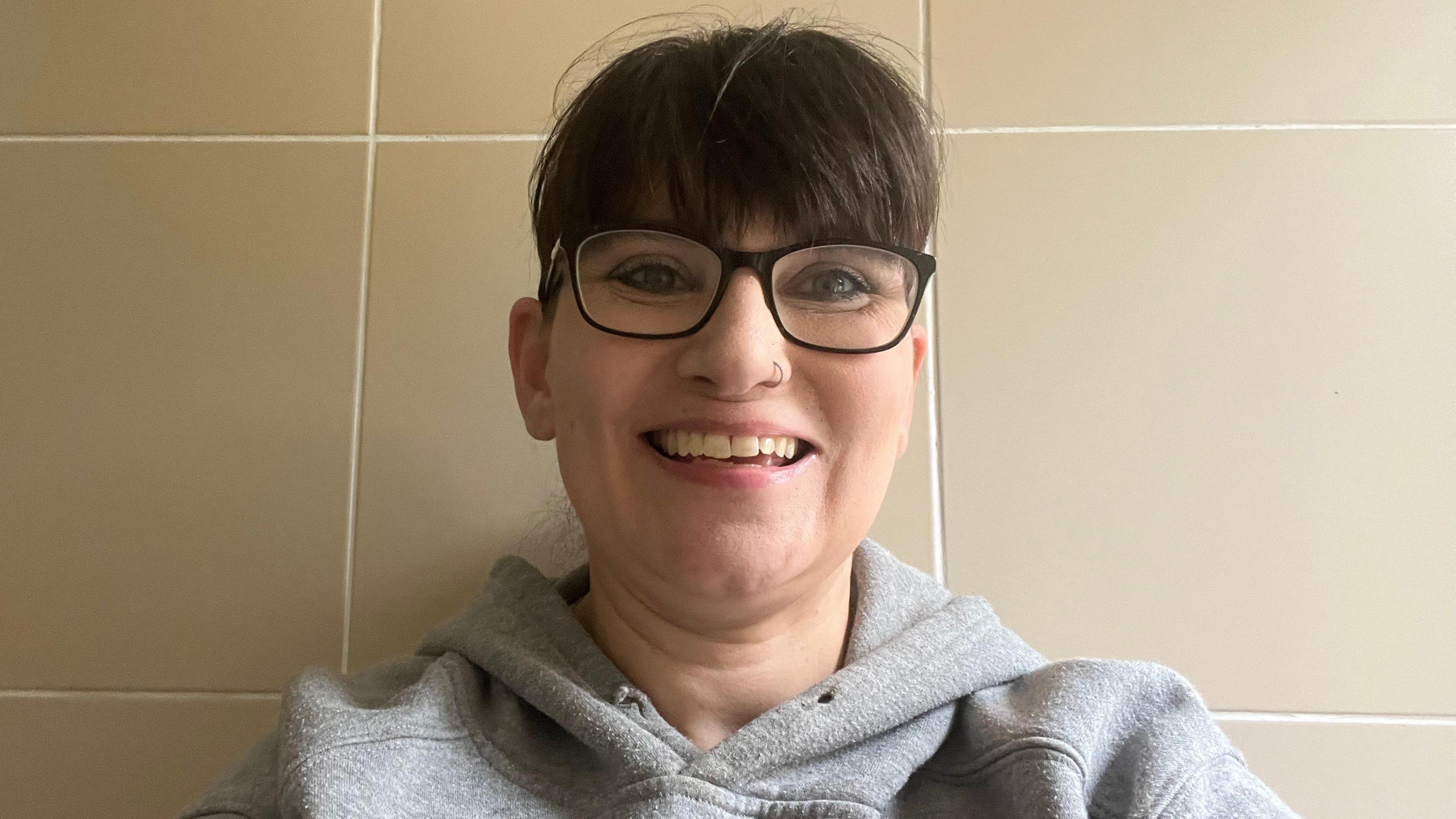 A woman with dark reading glasses and dark hair smiles into the camera. She is wearing a grey hooded top, and has a nose ring.