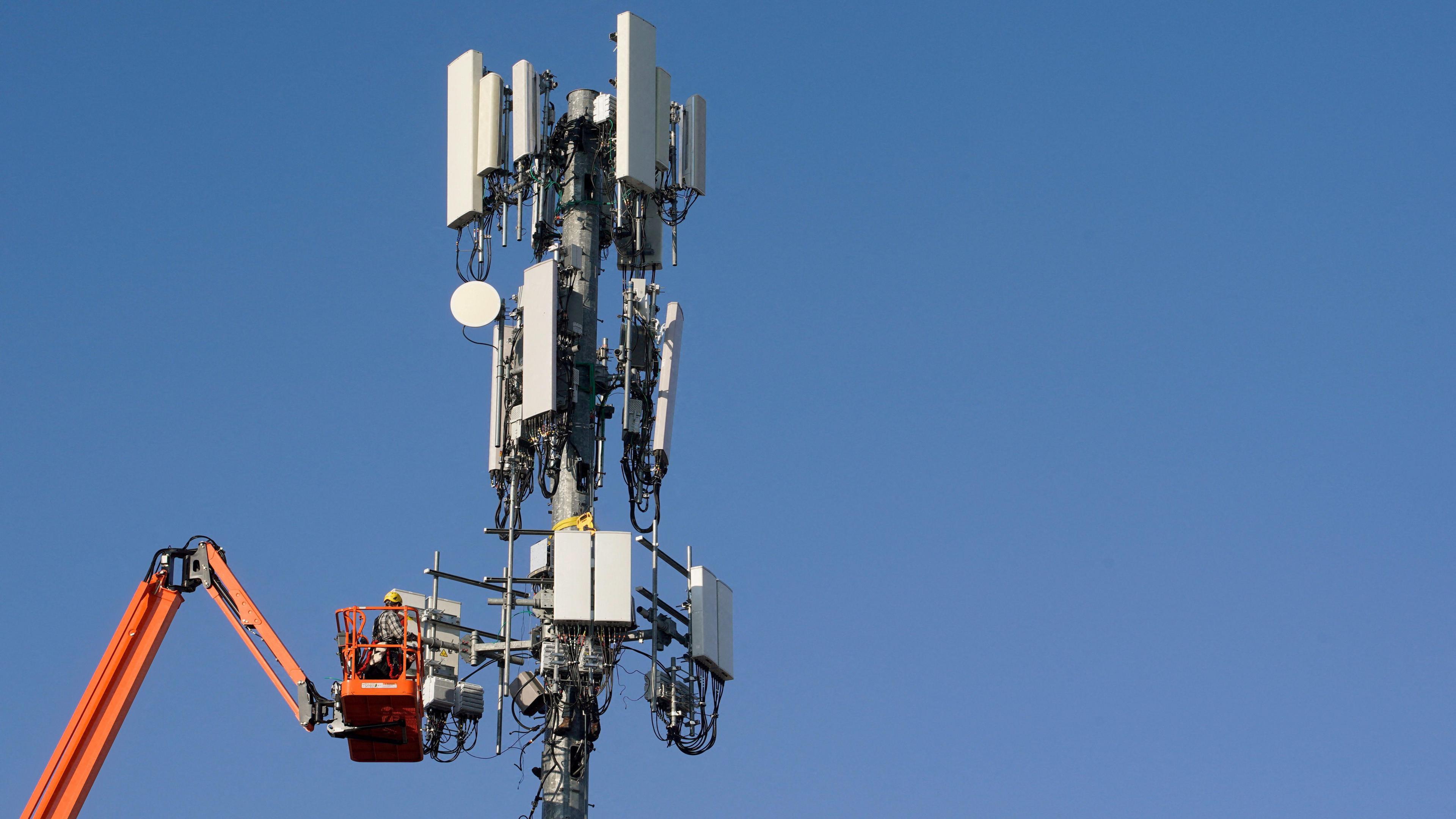 A phone mast in front of a cloudless sky, with an orange maintained crane working on it.