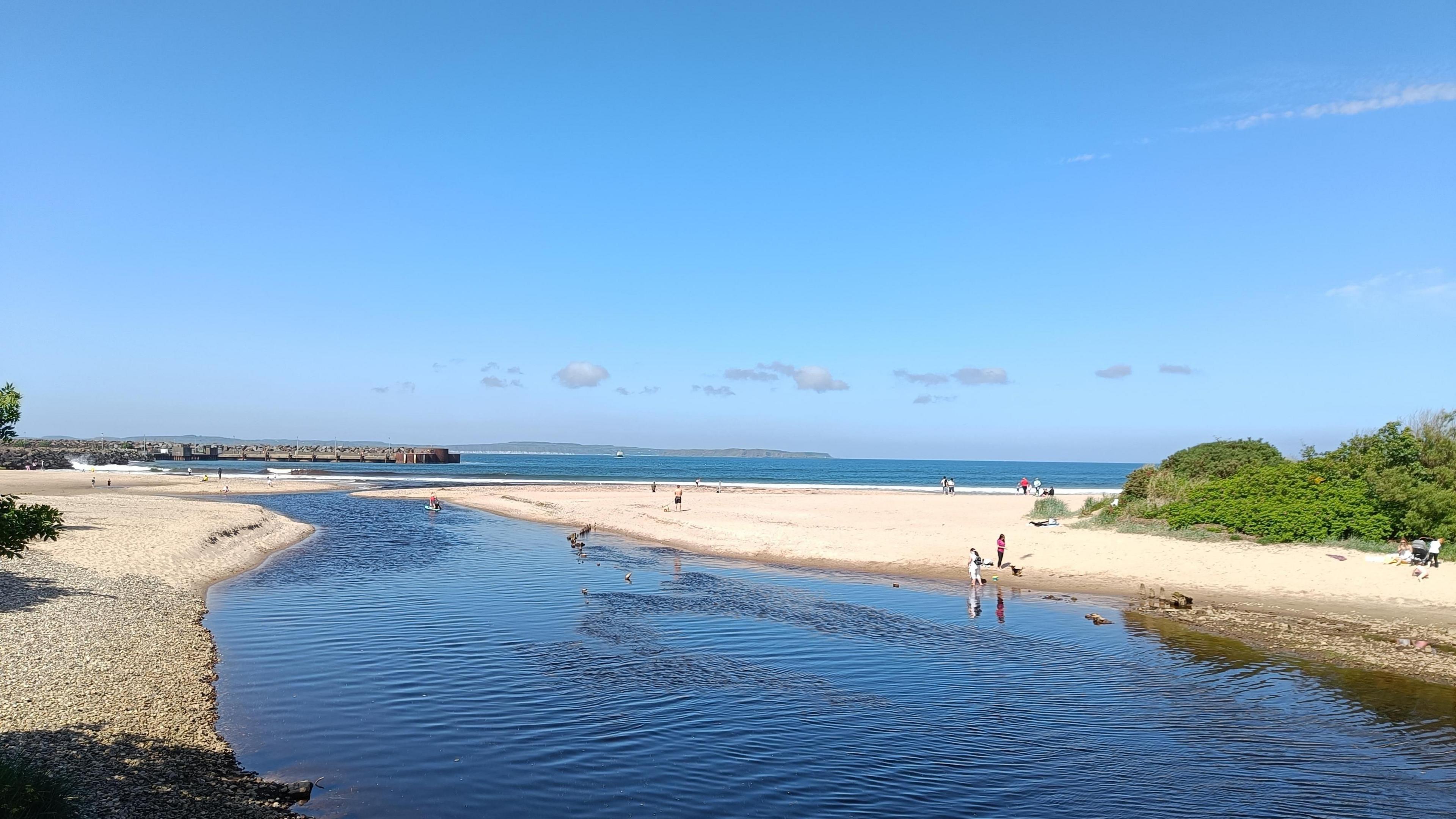 A river flowing into the sea with people on a beach and blue sky overhead