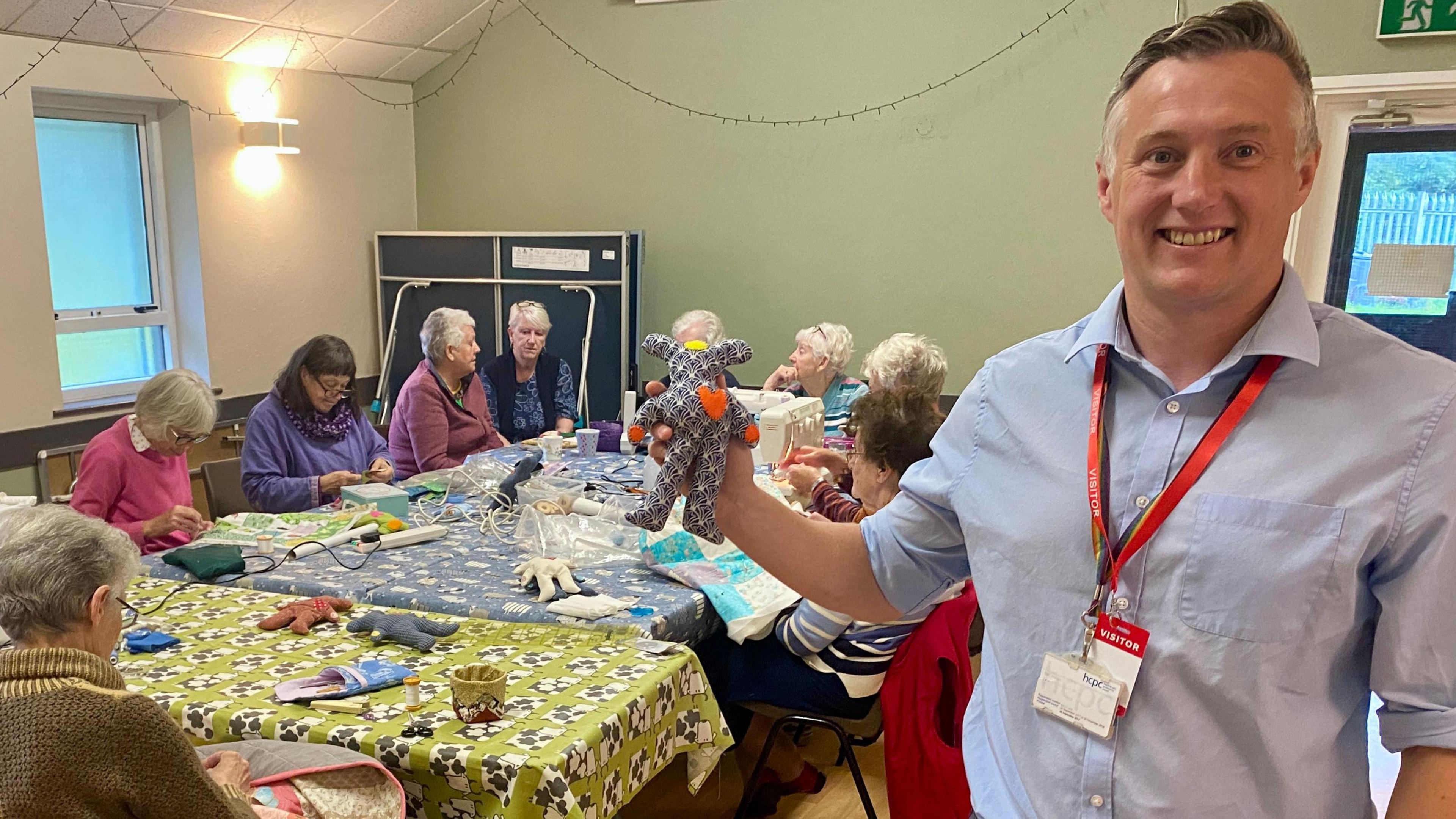 Alex Halsey is standing in a room holding a patterned sensory bear in his right hand which has an orange heart on it. Behind him is a table which ten women are sitting around. The women are stitching and chatting around the table which is covered with fabrics, sewing tools and sensory bears in various stages of creation.