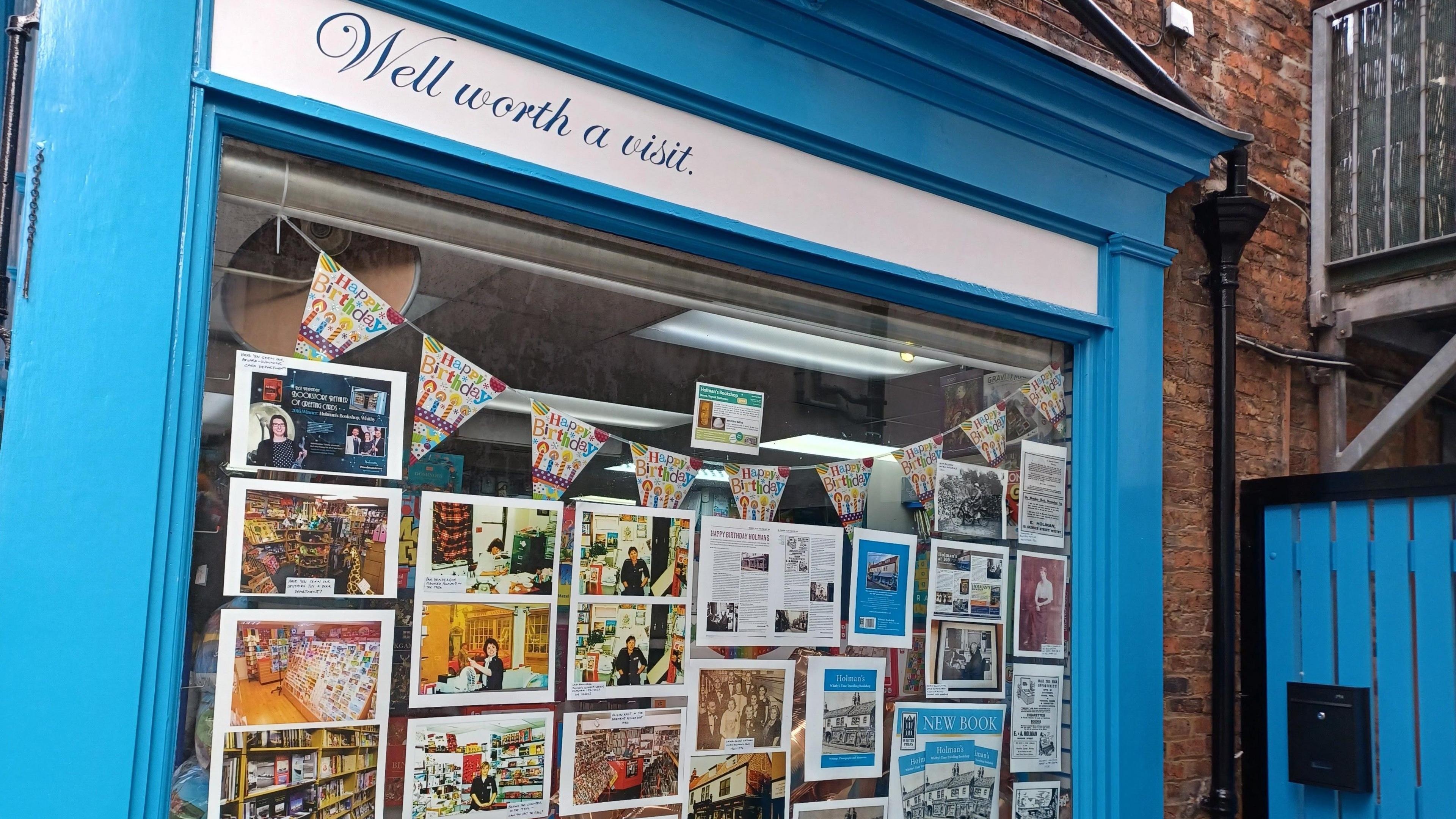 A shop window framed in bright blue paint with the words ‘Well worth a visit’ displayed at the top. The window features a collage of photographs, articles, and printed materials showcasing the store’s history and events.