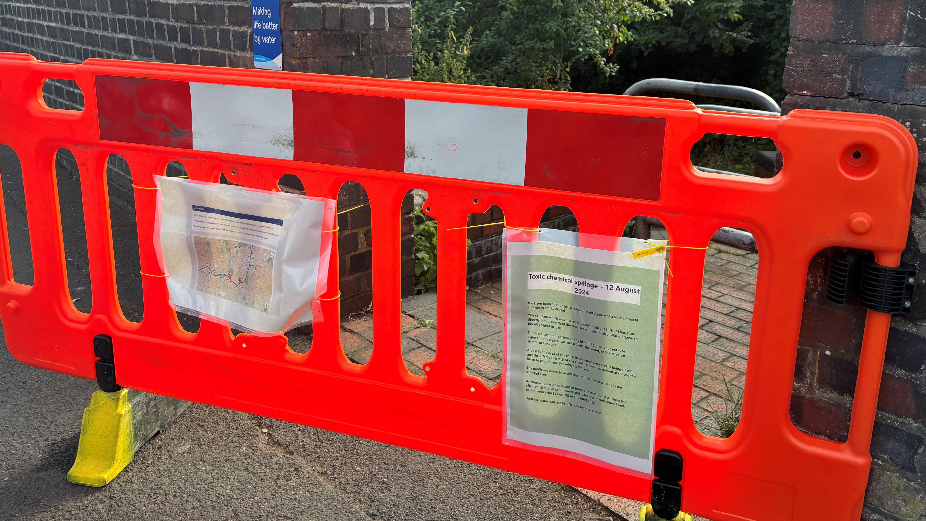 An orange barrier with two laminated signs attached, partially blocking a gap in a brick wall