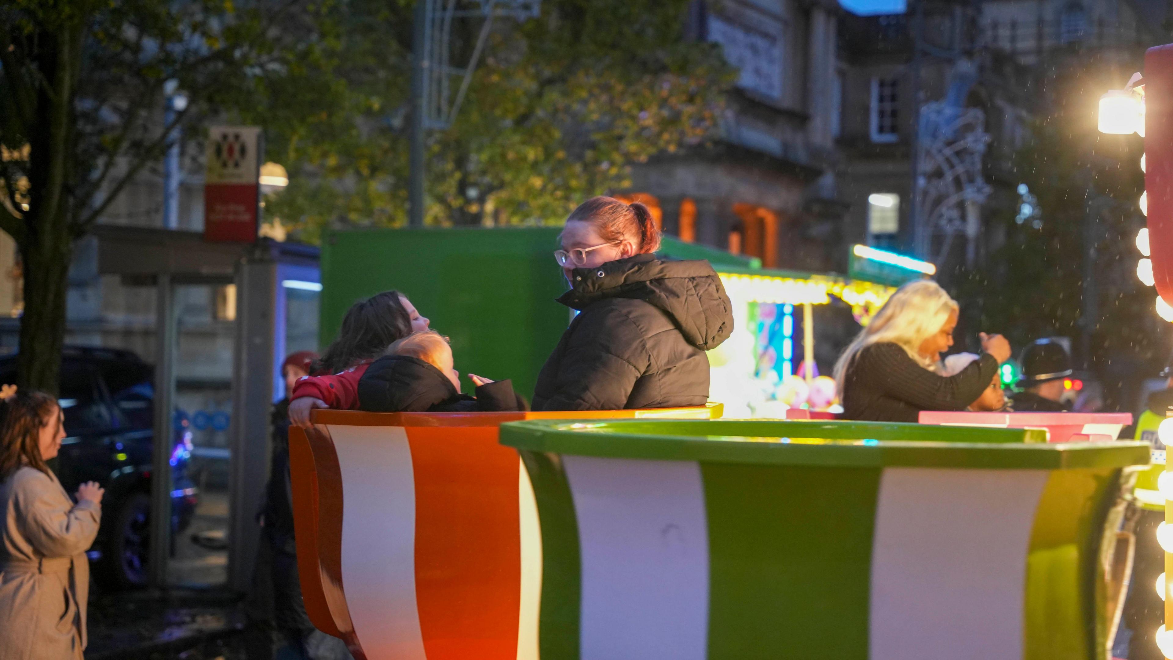 A woman with a ponytail sits with two children in a teacup right at the funfair held alongside the city centre switch-on. The teacups have red and white stripes and green and white stripes and the town centre can be seen in the background.