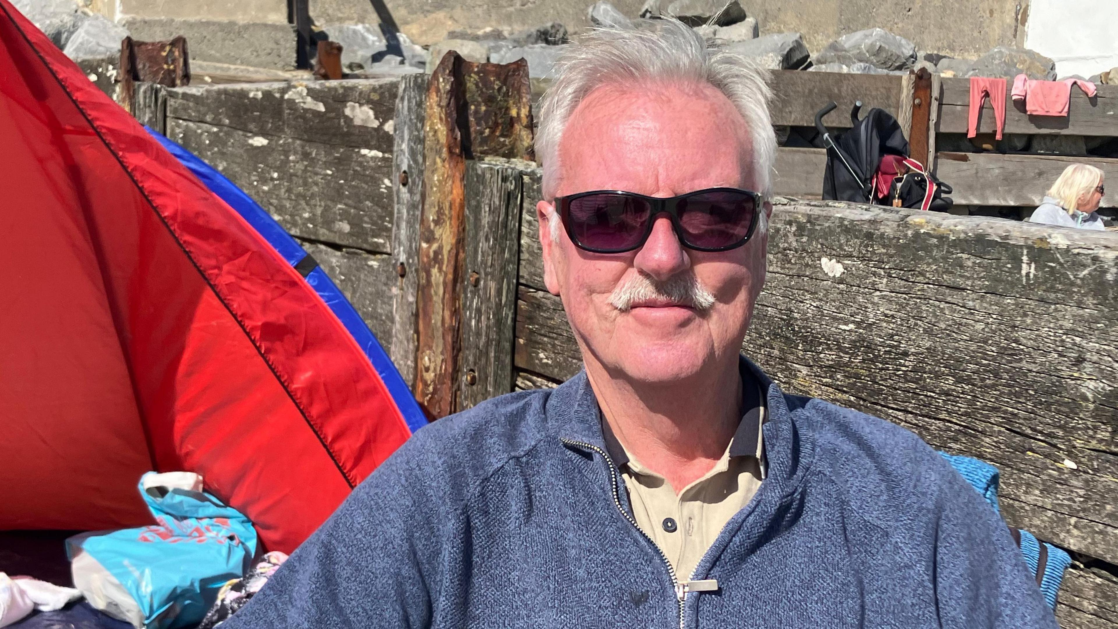 Allan Lunt sitting on the beach in a zip up fleece, sunglasses and white hair and moustache  