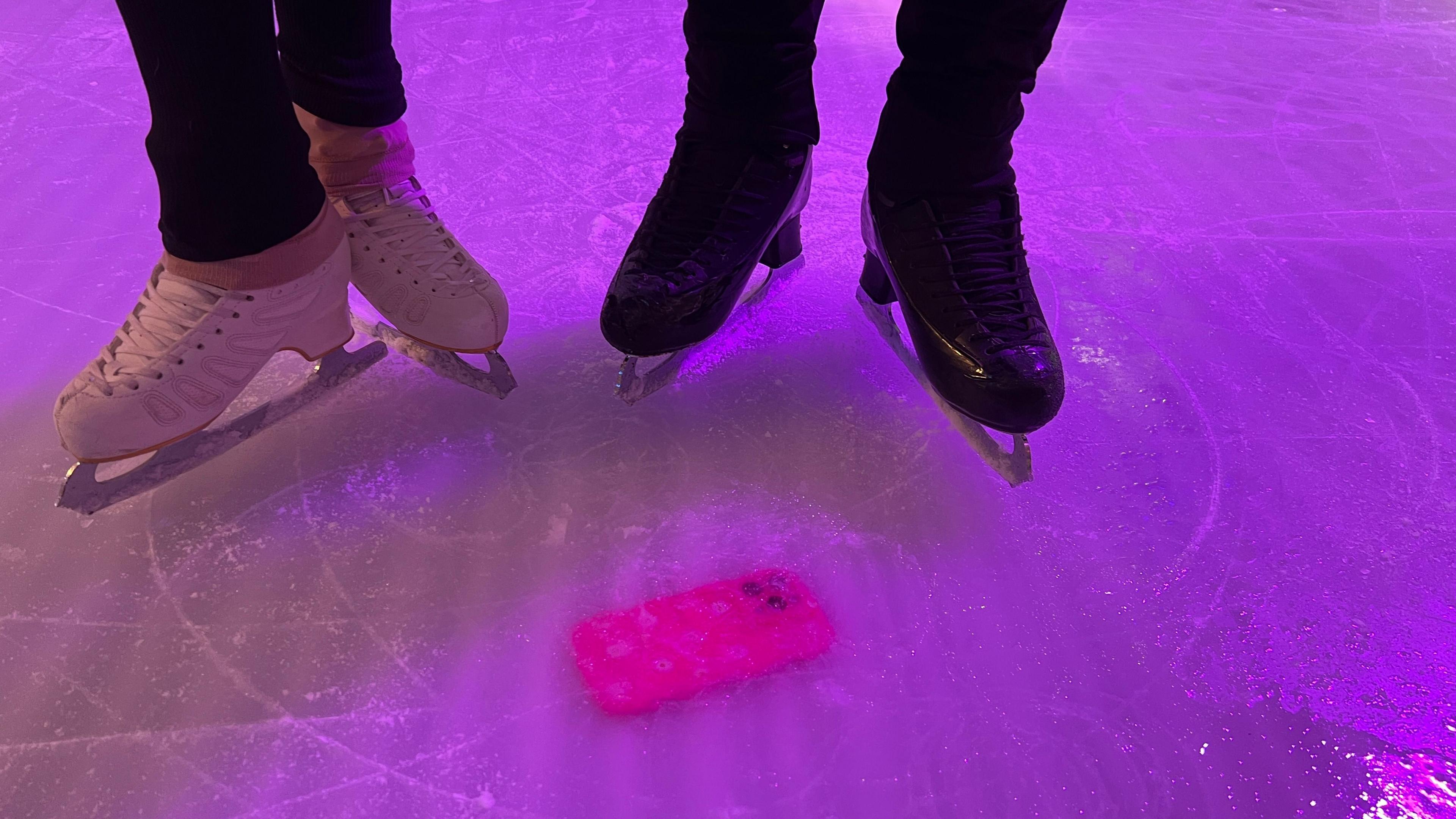 Bright pink phone case in ice. Standing next to it are the boots of two ice skaters. One set of white skates, to the left, and one set of black skates, to the right. 