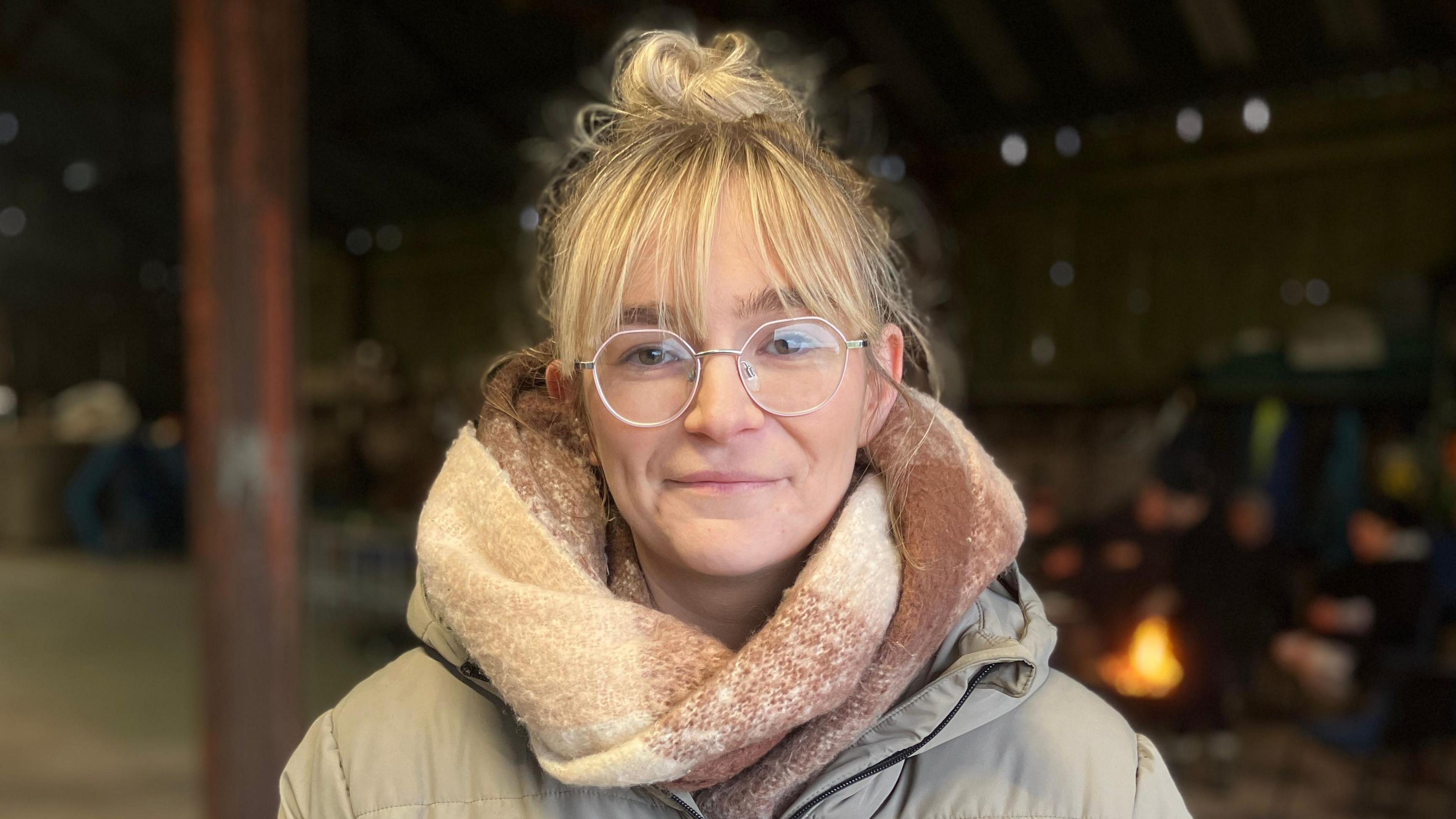 Sophie is looking at the camera smiling. She has blonde hair and is wearing glasses and a big cosy scarf in neutral tones. Out of focus over her shoulder women are warming their hands on an open fire. 