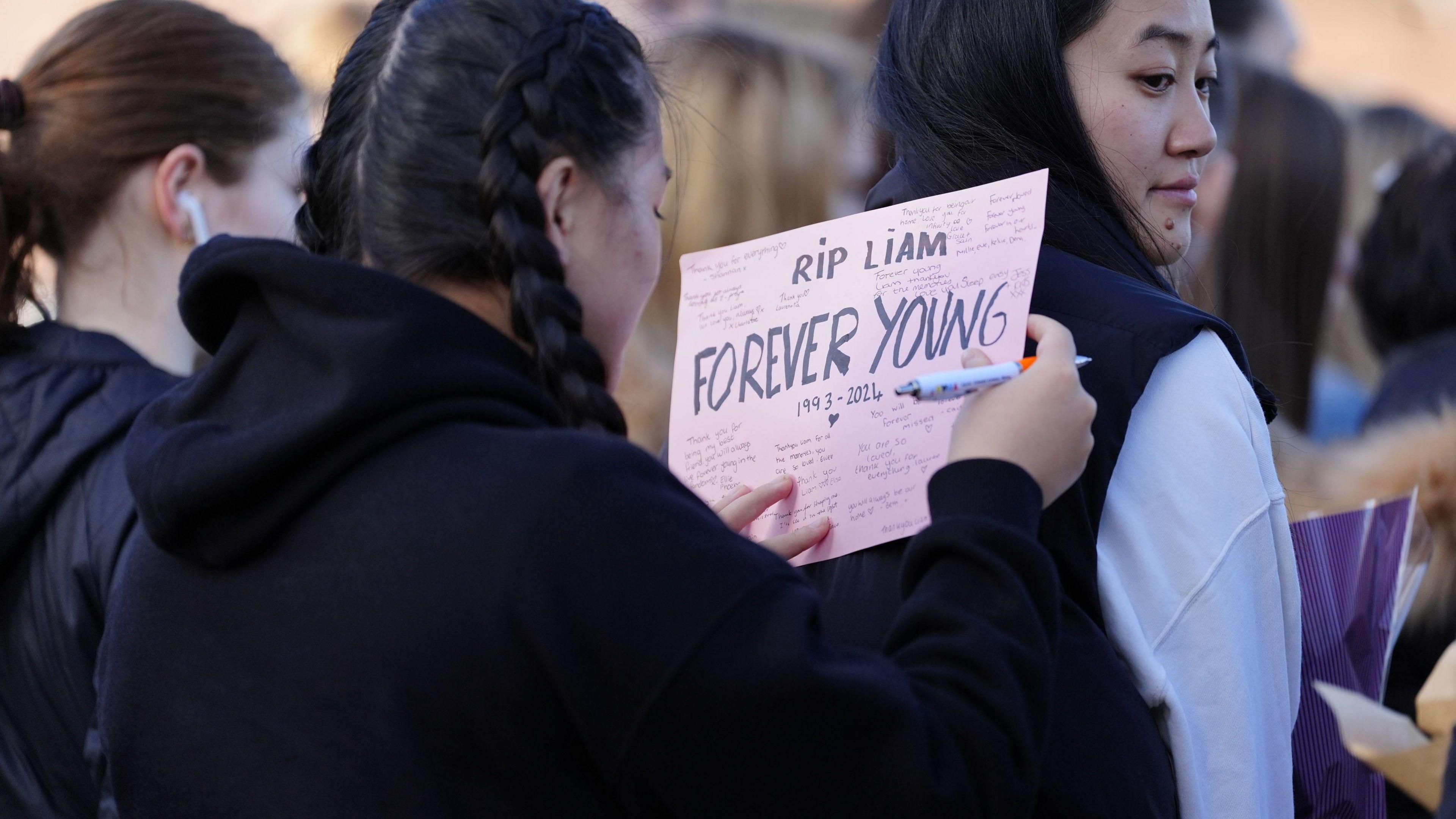 A young woman, with her black hair tied into plaits, writes on a piece of pink card which contains a number of messages as well as the words "RIP Liam" and "Forever Young". She steadies the paper on the back of another woman as she writes.