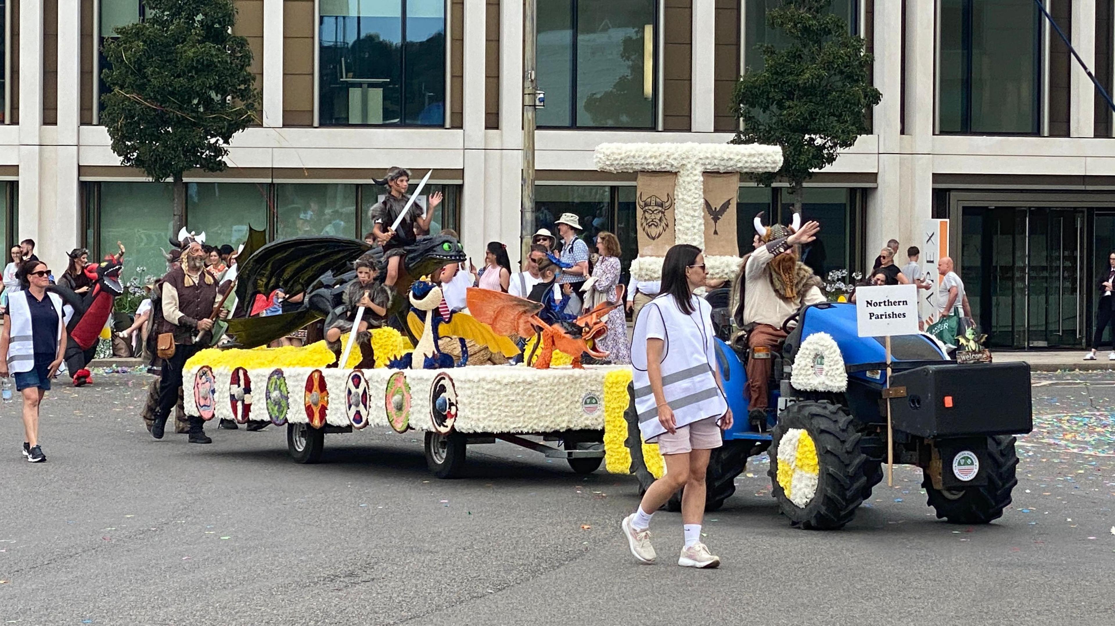 A float depicting a Viking boat. It is being towed by a small tractor and the driver is wearing a Viking helmet and long orange beard. A sign on the tractor reads NORTHERN PARISHES. On the float there are boys holding oars wearing dressed as Vikings. The side of the float has seven different shields. Stewards walk alongside the float.