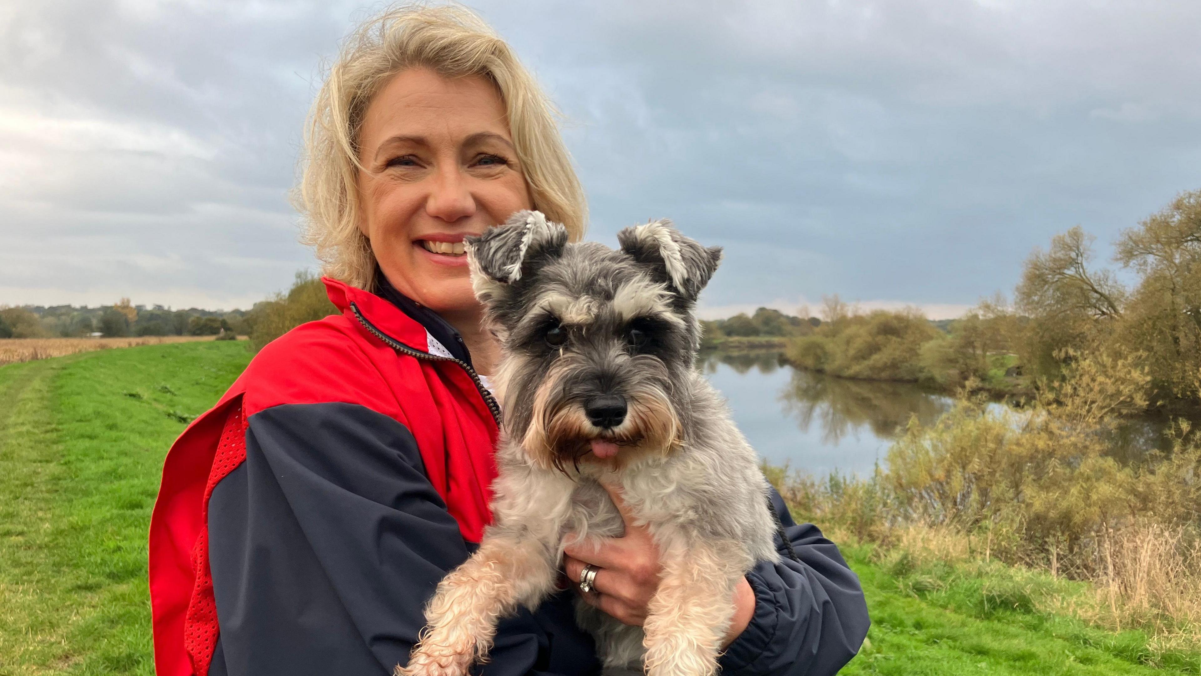 Blonde-haired woman holding a dog next to a river