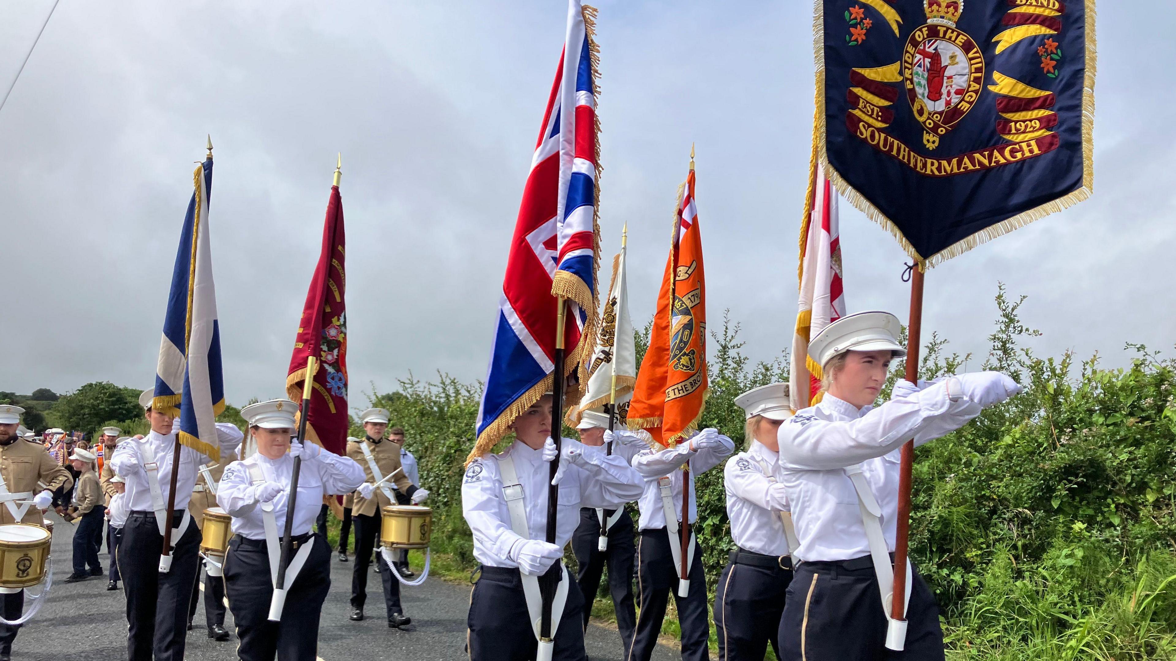 Women marching in white shirts and white flat top hats. They each carry a flag or banner, the front banner is black and says South Armagh on it, there is also a Union Jack flag. They are followed by men with marching drums