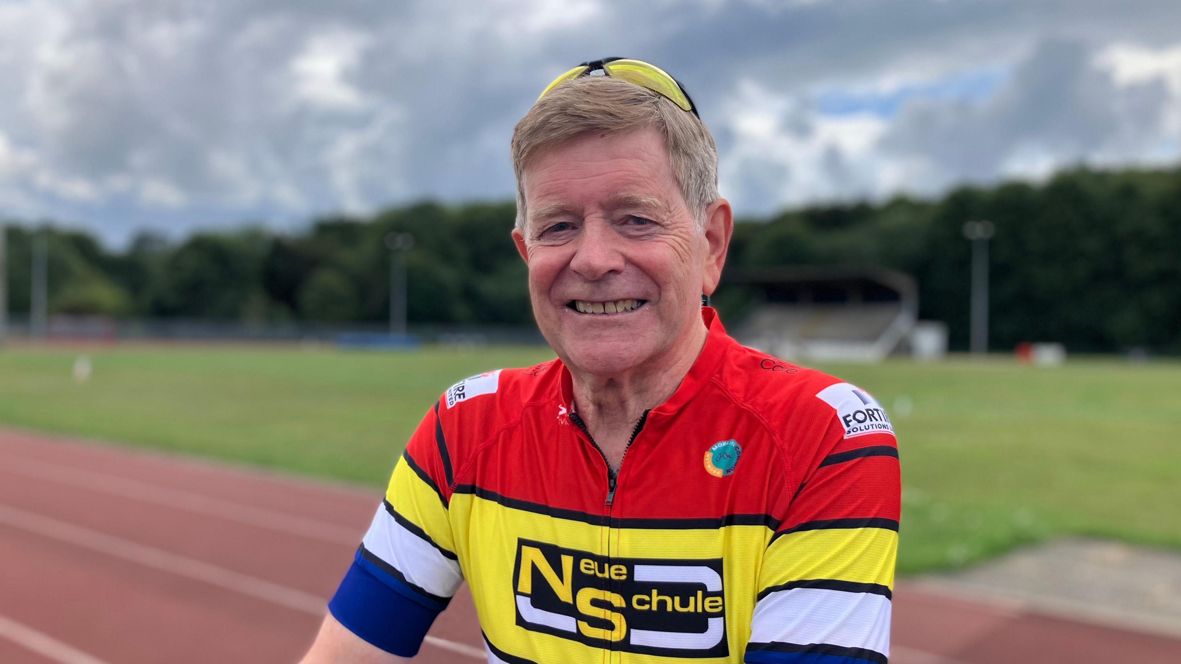 A man with grey hair smiles at the camera. He wears yellow sunglasses on top of his head and a red, yellow, white and blue cycling jersey. Behind him, a race track is blurred.