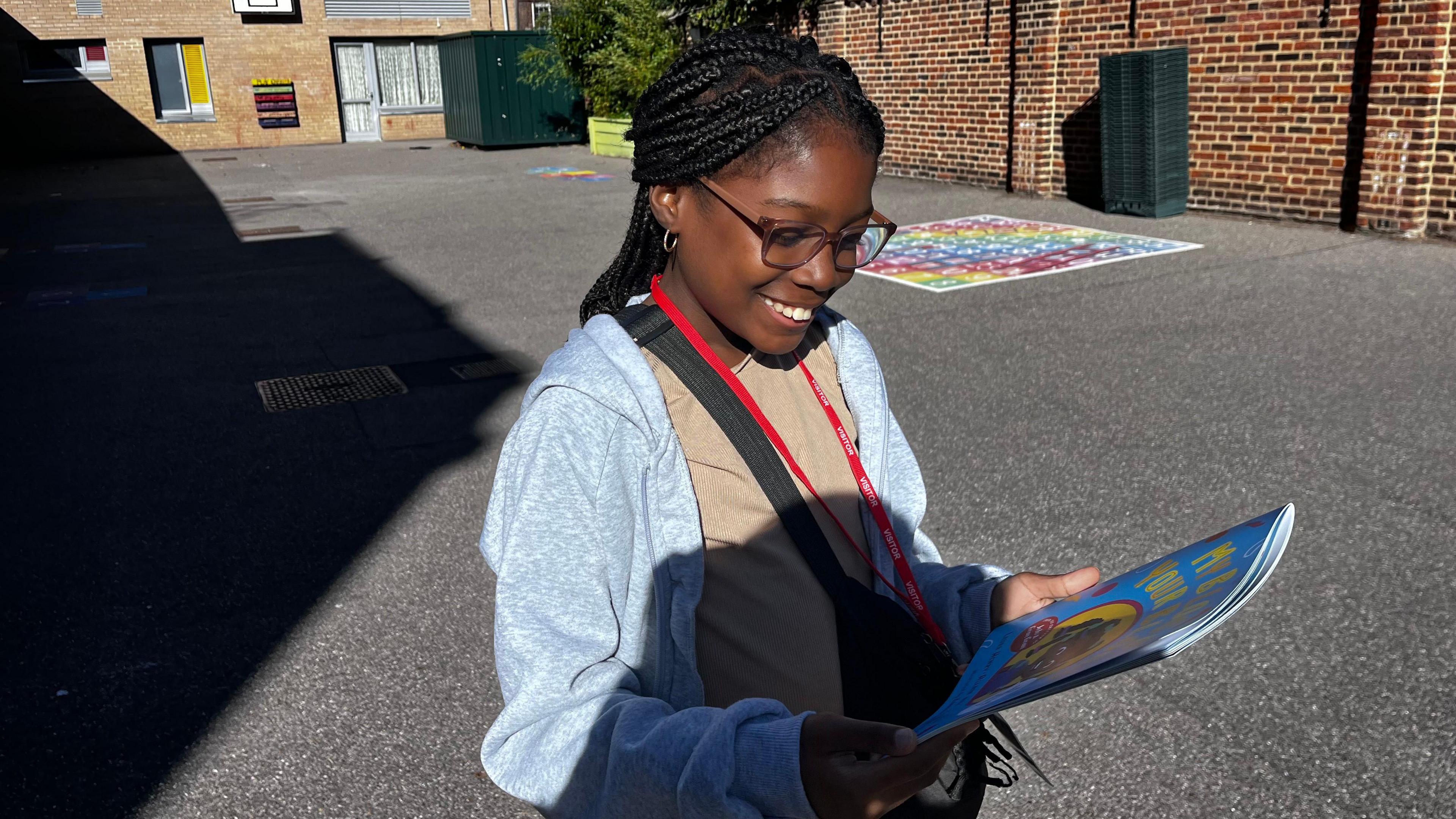 A young girl, Angel, standing in a school playground looking down at a copy of the book, 'My Blood, Your Blood'. She is smiling. She is wearing glasses, has dark skin, and has black braided hair. She is wearing a sand-coloured top, a grey jumper, and a red lanyard.
