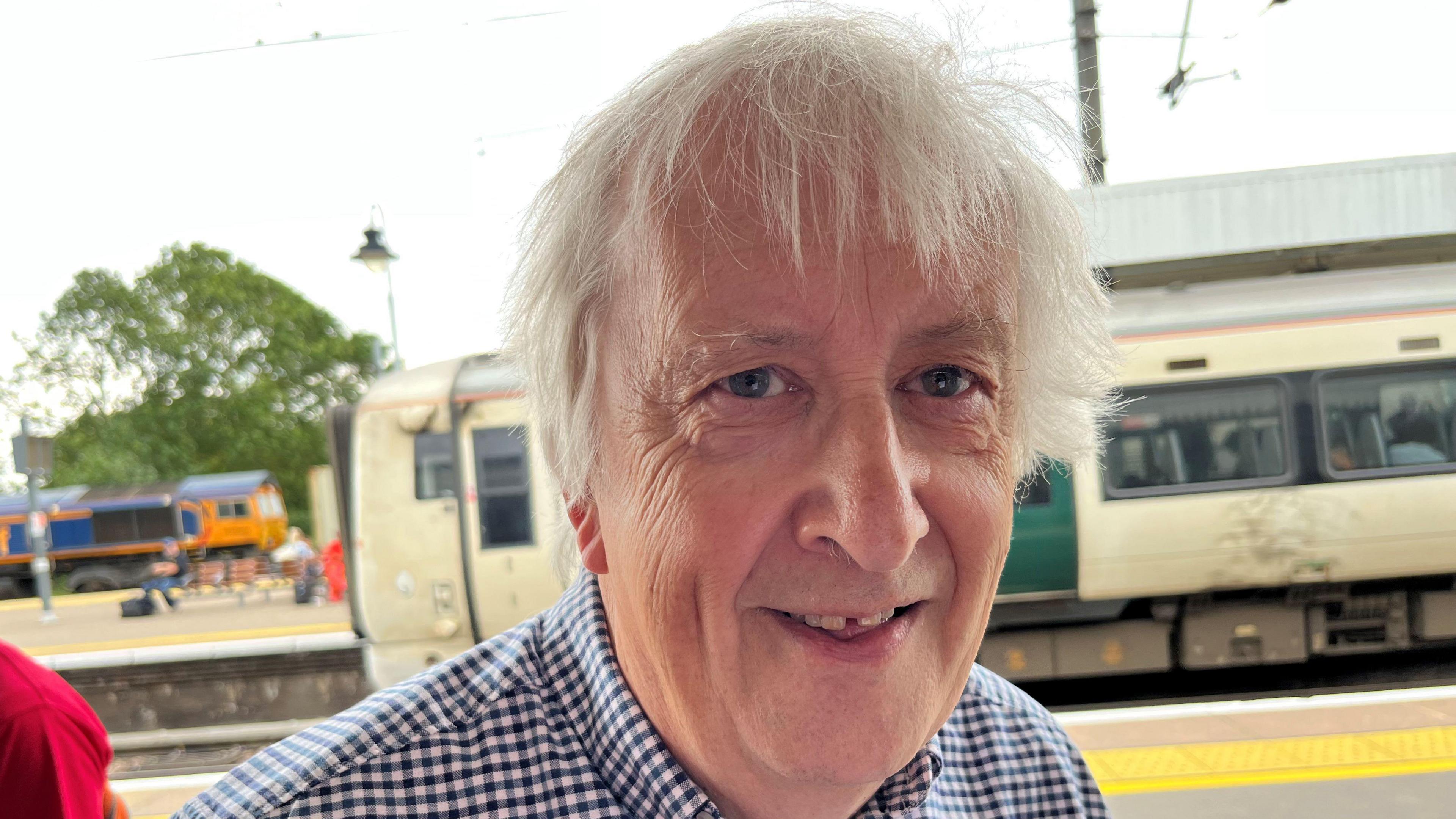 Geraint Hughes has quite long white/grey hair over his forehead with a fringe over his eyes, he is wearing a black and white checked shirt and behind is a train at the platform.