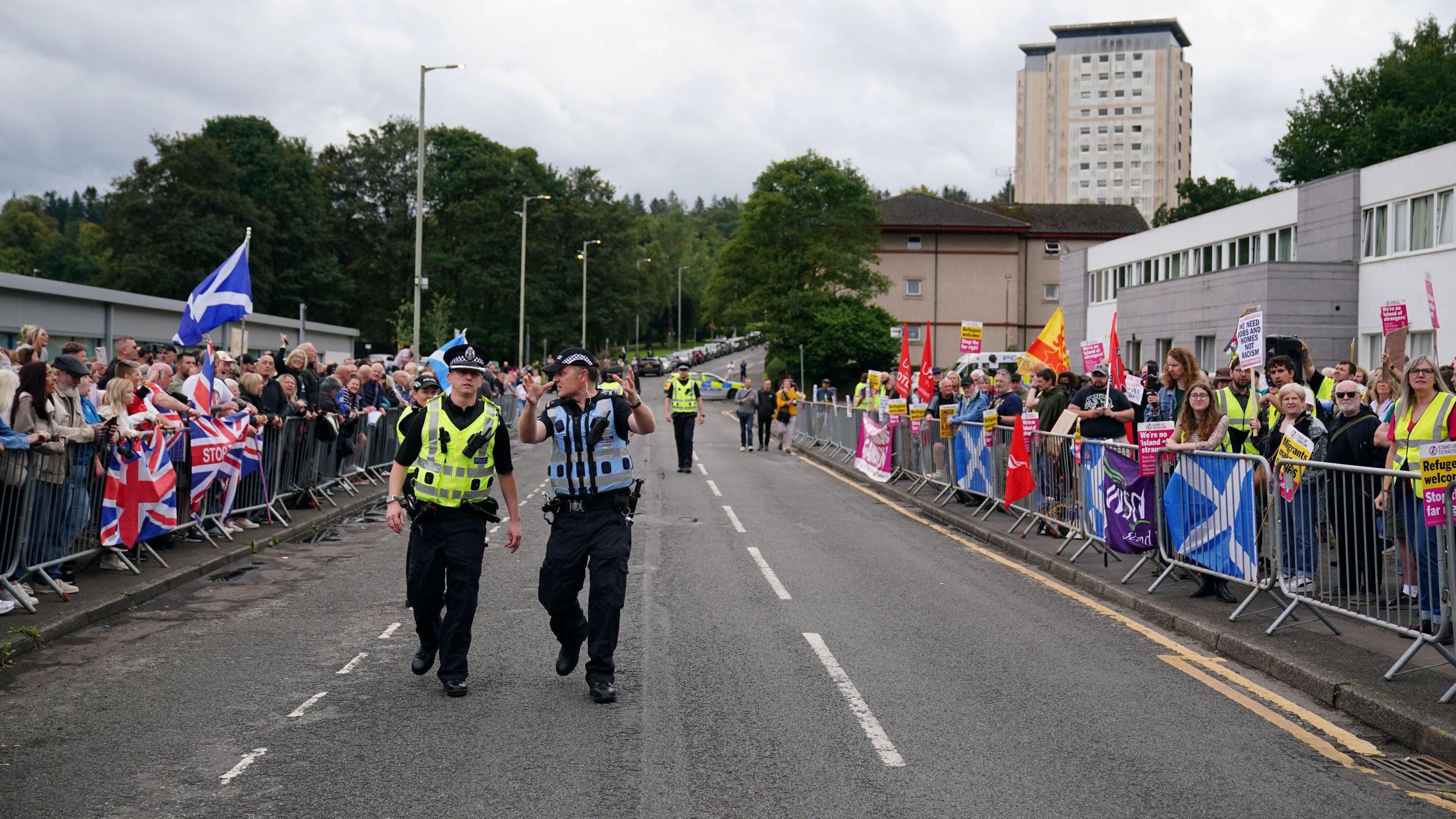 Two police officers walk down an empty road. One of the officers is turning to talk to the other. Either side of the road are opposing groups of protesters, and they wave signs with slogans and Union and Saltire flags. A tower block dominates the skyline.