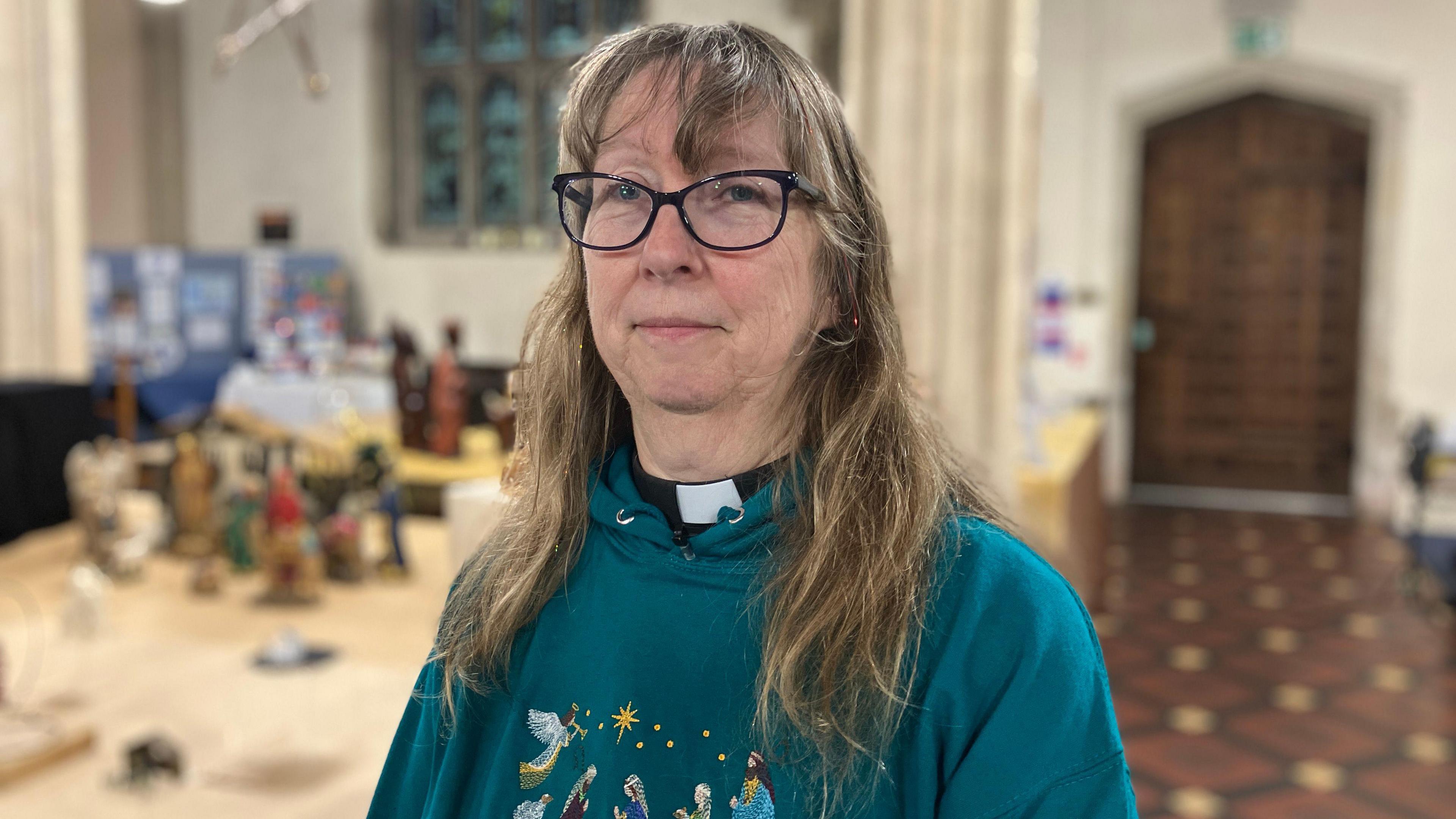 Rev Chris Butler. She has long hair and is wearing black framed glasses and a green hoodie with a nativity scene embroidered on the chest. She is looking directly at the camera and smiling. Some of the nativity displays can be seen out of focus, in the background behind her.