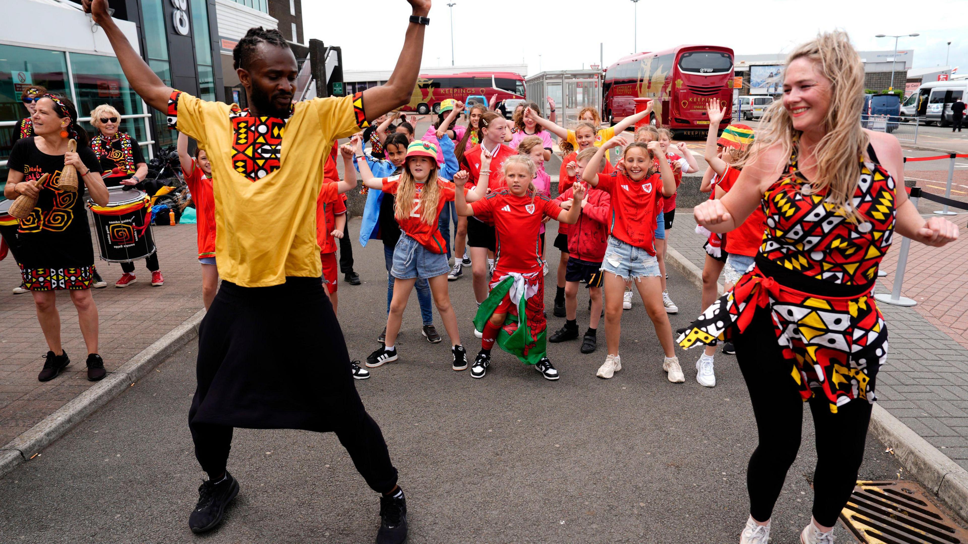 A man wearing a yellow shirt and dark trousers dances with a woman wearing a multi-coloured top. A group of children are also dancing behind them.