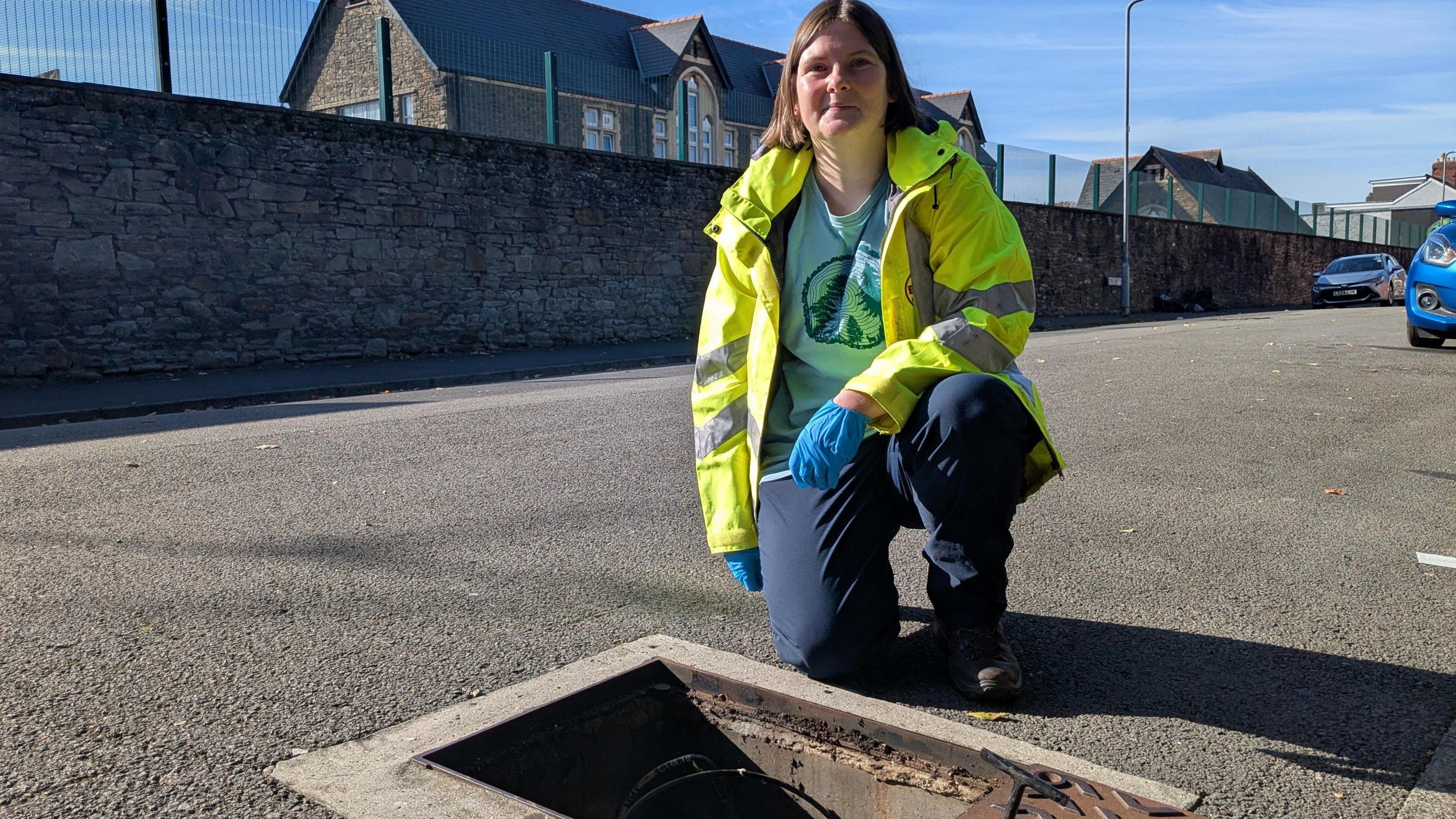 A women with brown hair kneels next to an open drain cover on a quiet residential road. She's wearing a high vis jacket and blue gloves