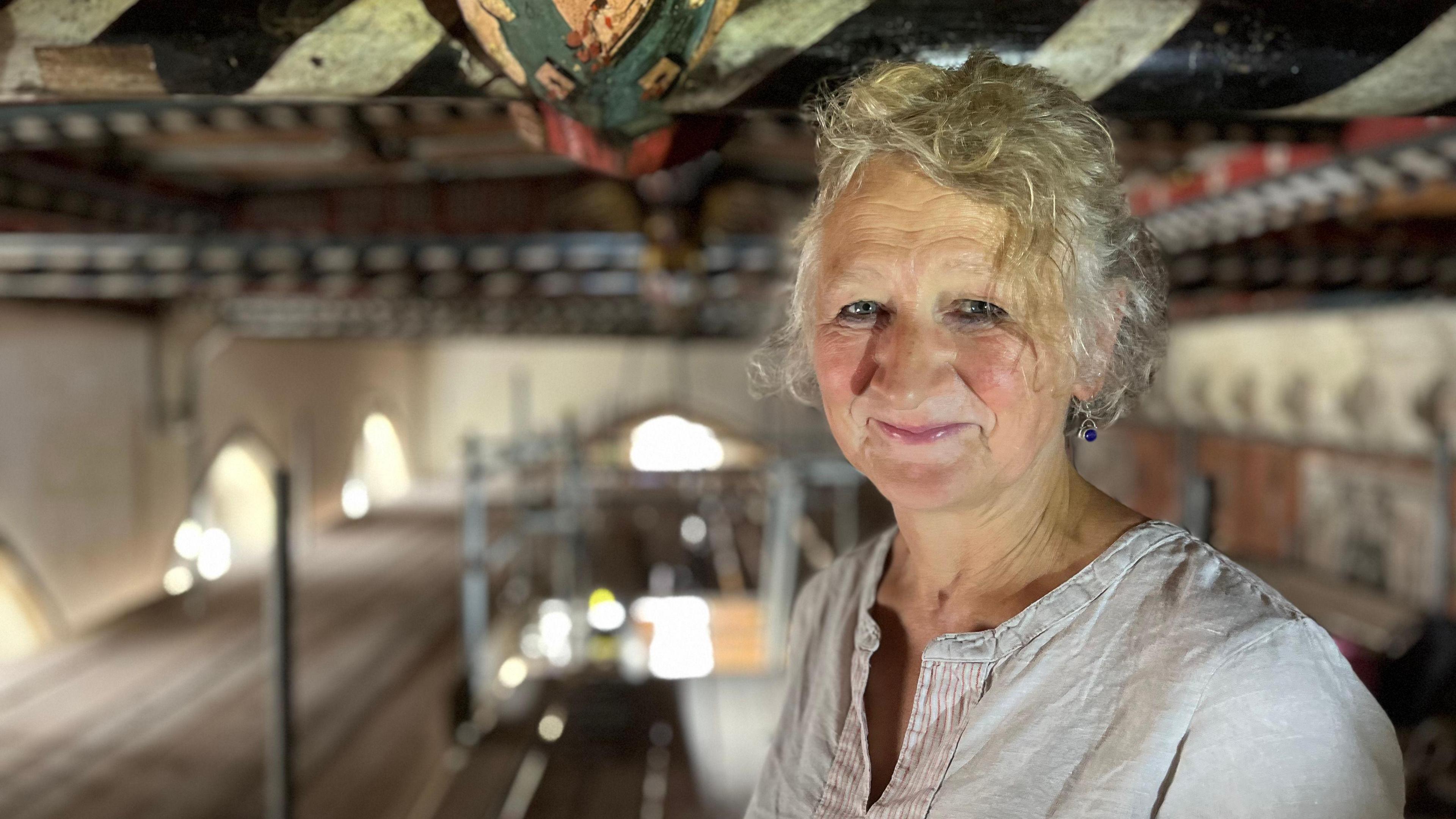 Ruth smiles at the camera. Her hair is tied up and she wears a light-coloured linen top. She stands in the ceiling area.