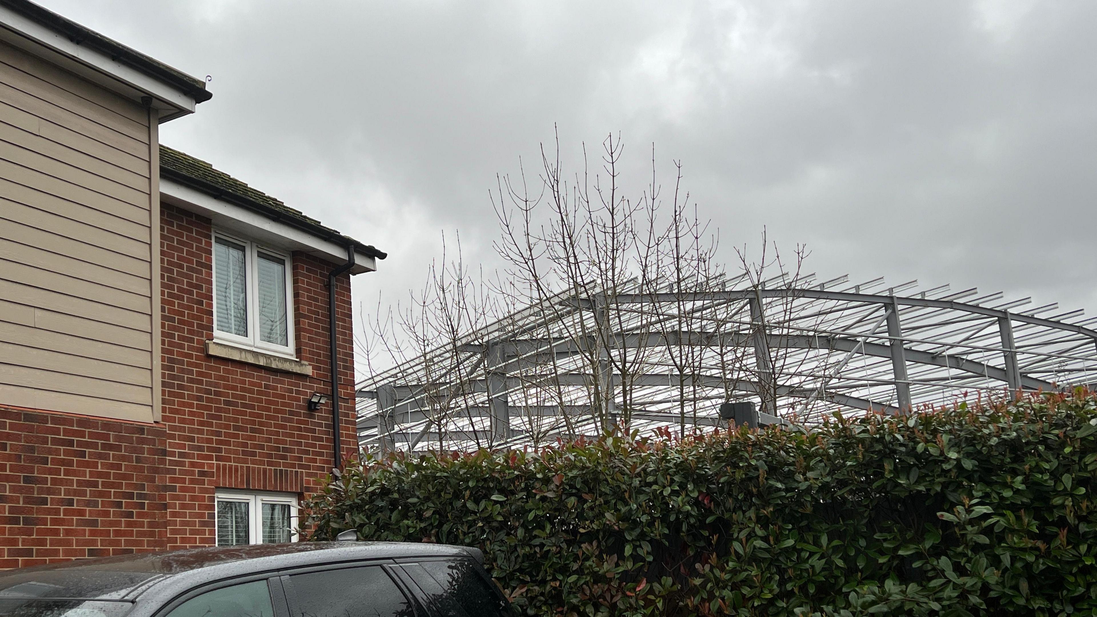 The corrugated metal structure of the warehouse can be seen looming over the top of a resident of Hooke Close's hedge next to their property. The resident's black car is also in shot by a red brick house.