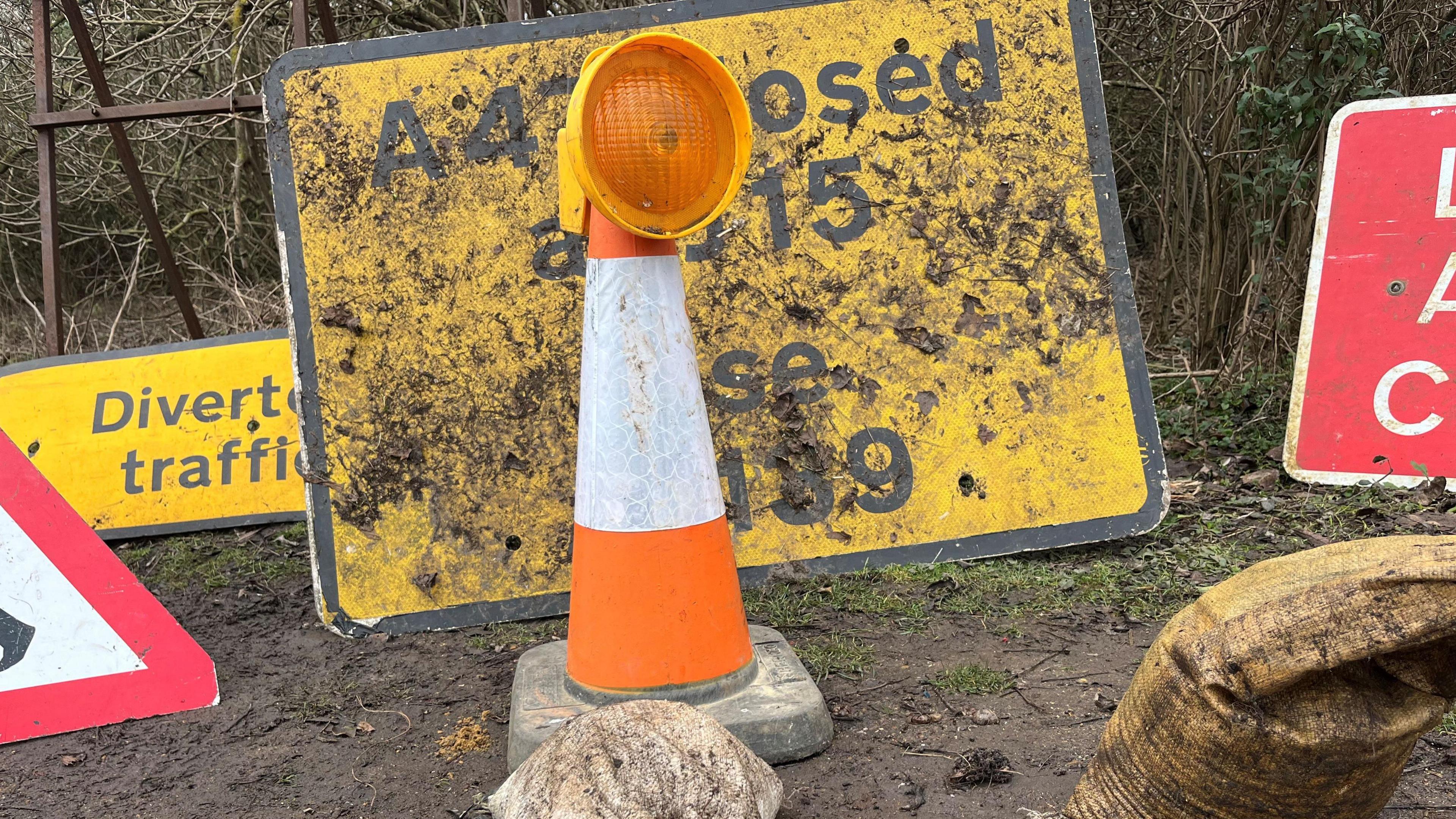 An orange and white traffic cone with a yellow safety flash lamp on top, around it are various road signs and sandbags, all found by Mr Fishpool on the verges and hedges along Peterborough's roadways.