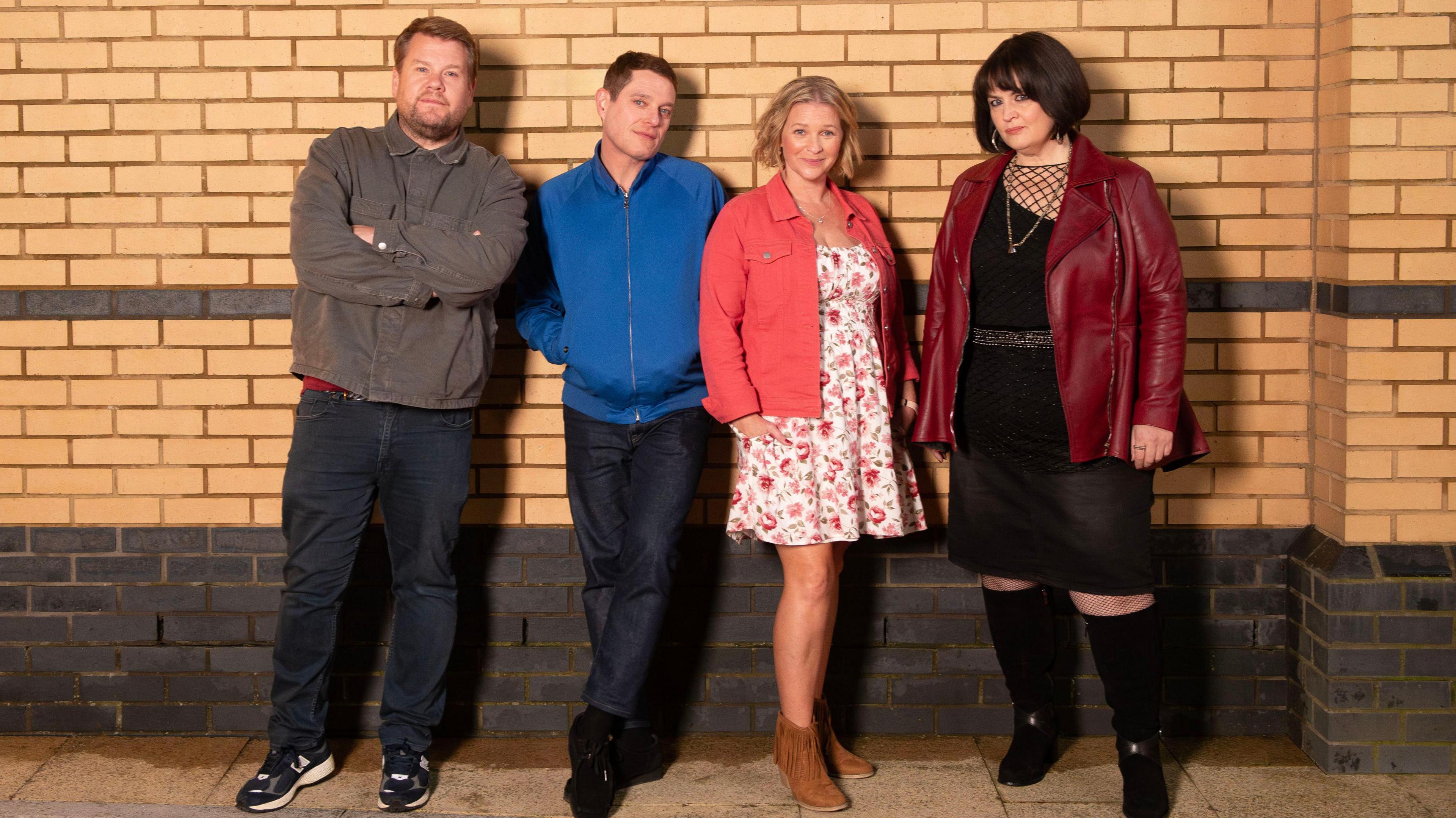 Four members of the cast of Gavin and Stacey reunite for an official cast photo: Smithy, Gavin, Stacey and Nessa. They are stood against a brick wall. Smithy (played by James Corden) is wearing a grey jacket and blue jeans and has his arms crossed, Gavin (played by Mathew Horne) is next to him, wearing a blue jacket with his hands in the pockets and blue jeans. Next to him is Stacey (played by Joanna Page), she is wearing brown cowboy boots, a white floral dress and a pink jacket, and she is stood next to Nessa (played by Ruth Jones), who is wearing a burgundy leather jacket and a black dress with long black boots.