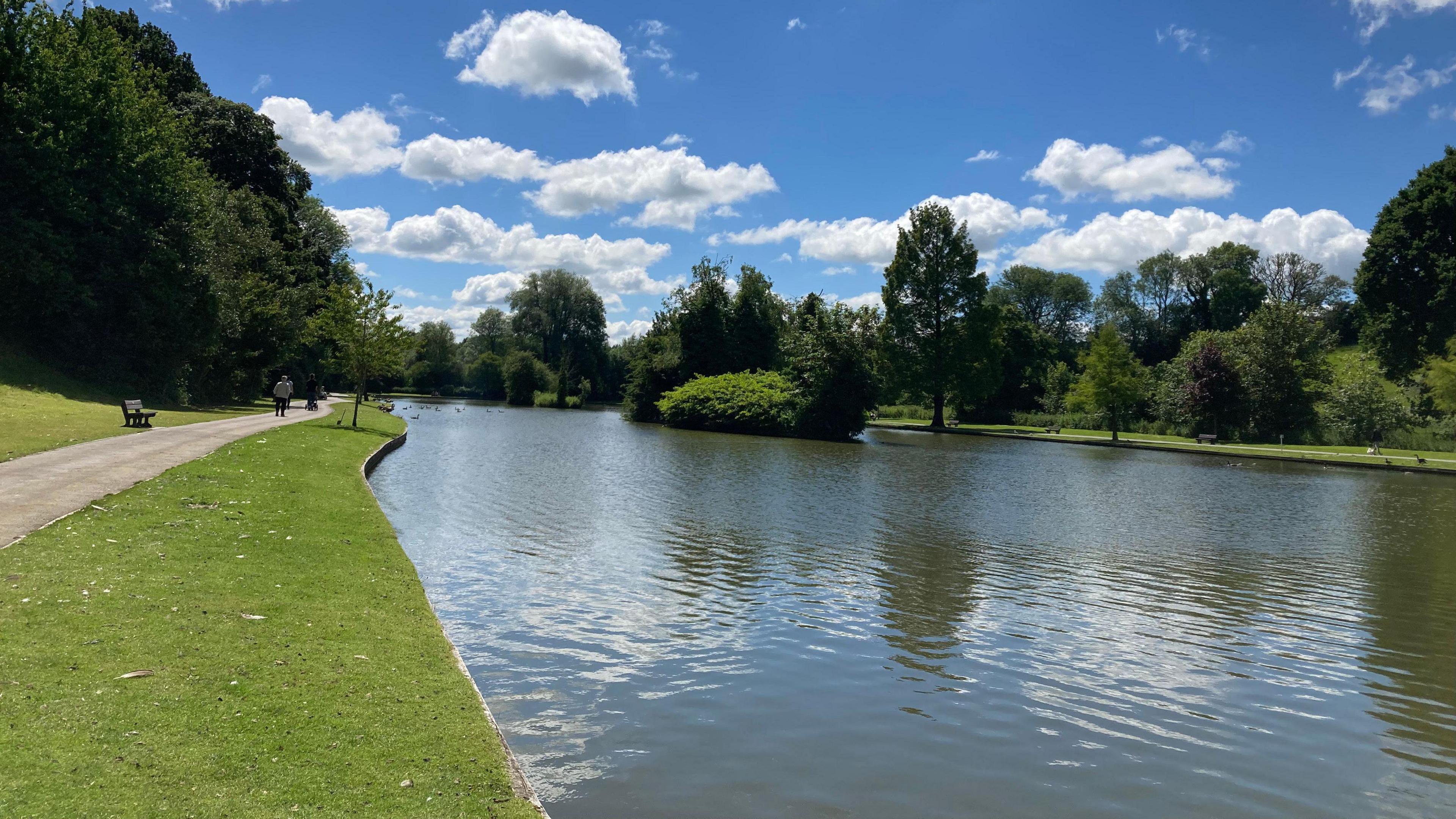 Lake with island in the middle, grassy banks with path and benches under a blue sky