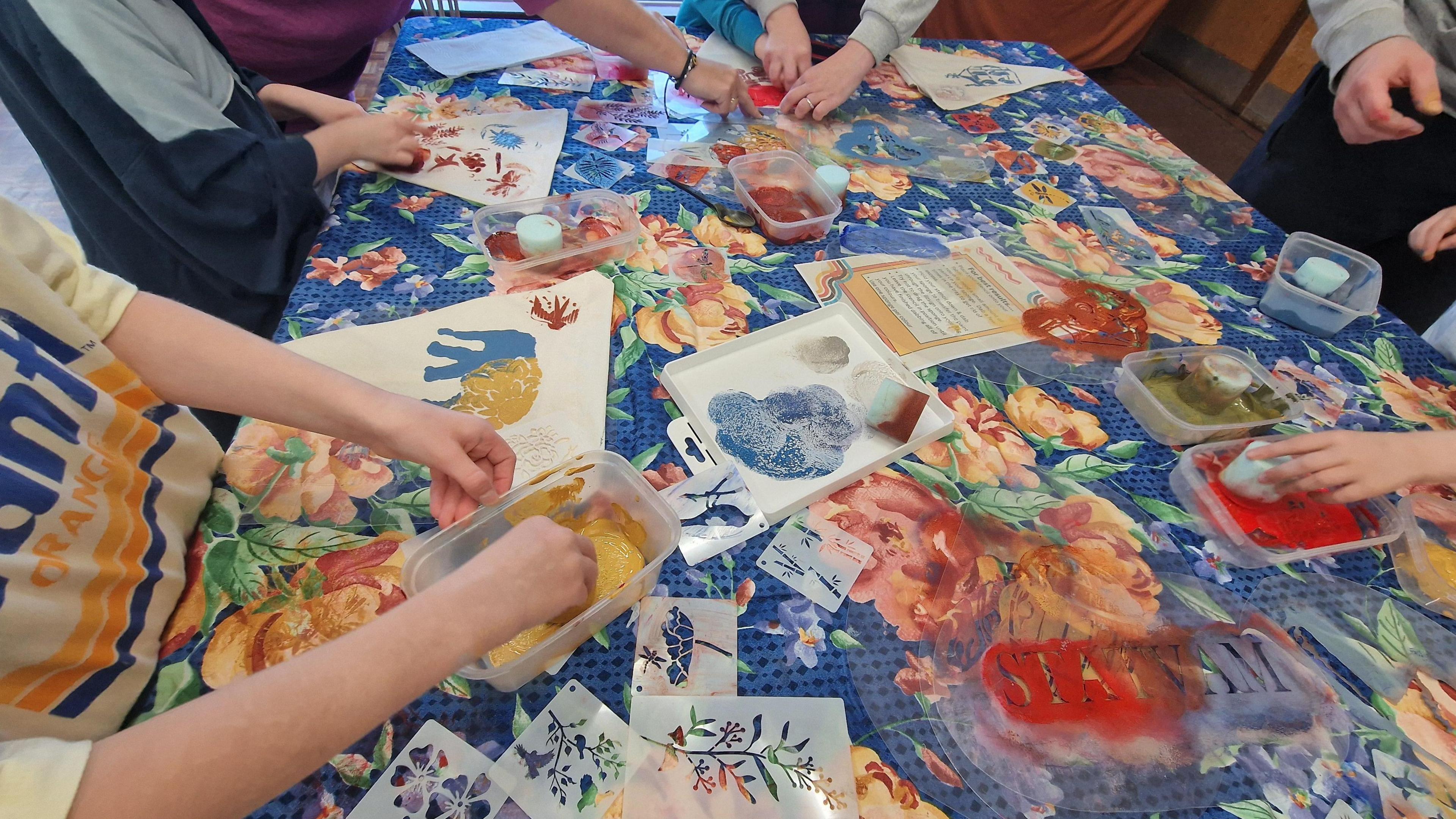 Children printing with paint on a table which is decorated with a florla tablecloth.