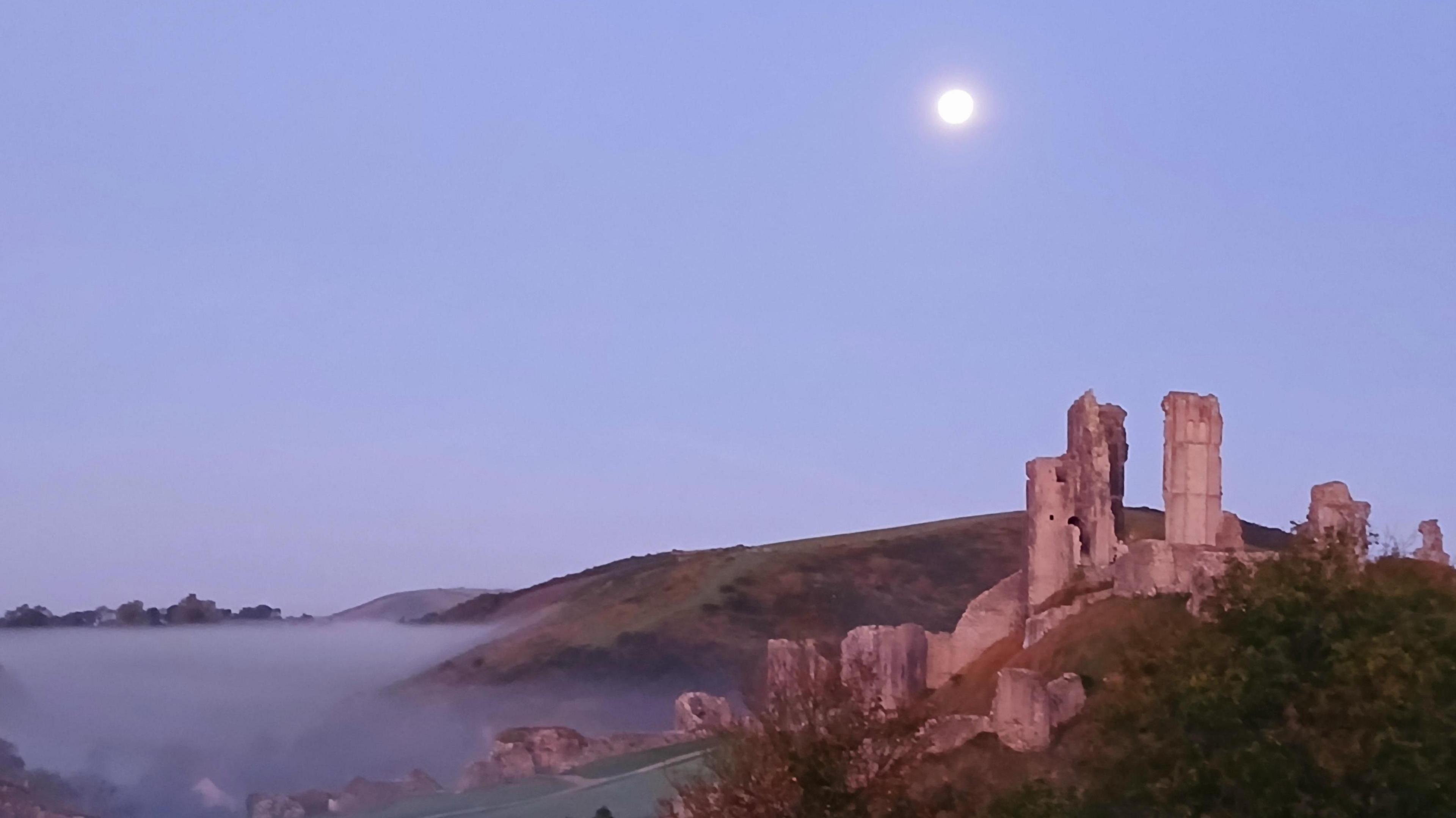 The moon appears as a bright circular light about the ruins of corfe castle. There are misty fields and bushes surrounding the castle