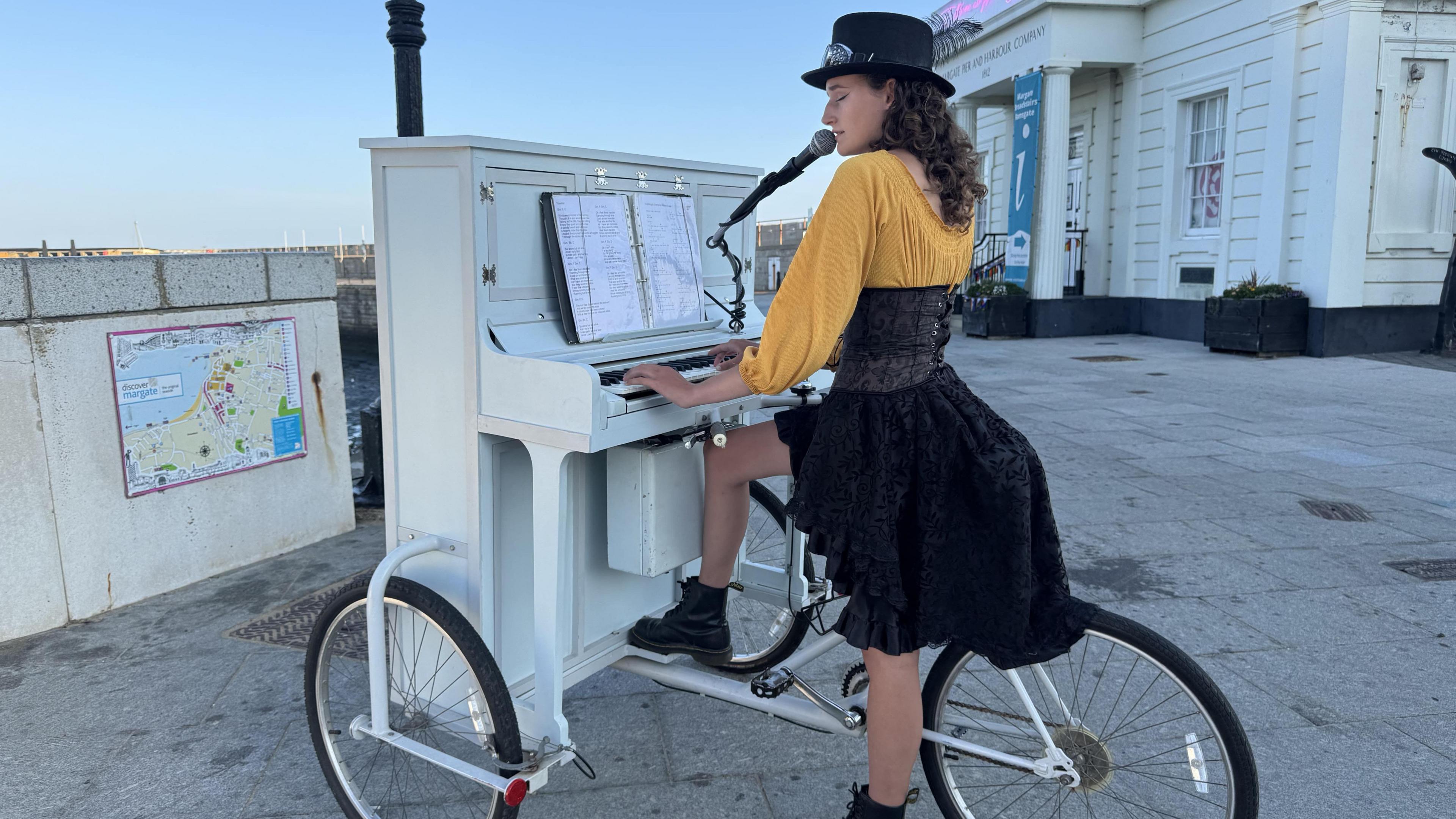 Chloe Marie Aston in a yellow cardigan and black dress stands sits on her bike while playing the piano.