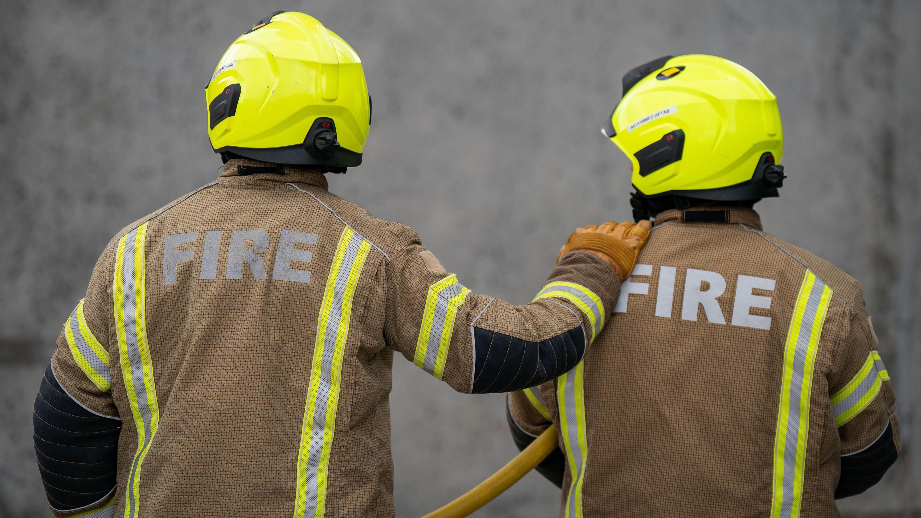 Two firefighters are wearing protective brown clothing with silver and yellow reflective strips. They have yellow helmets on their heads and one is wearing a brown glove with their hand on the other's shoulder. The other firefighter has a yellow hose