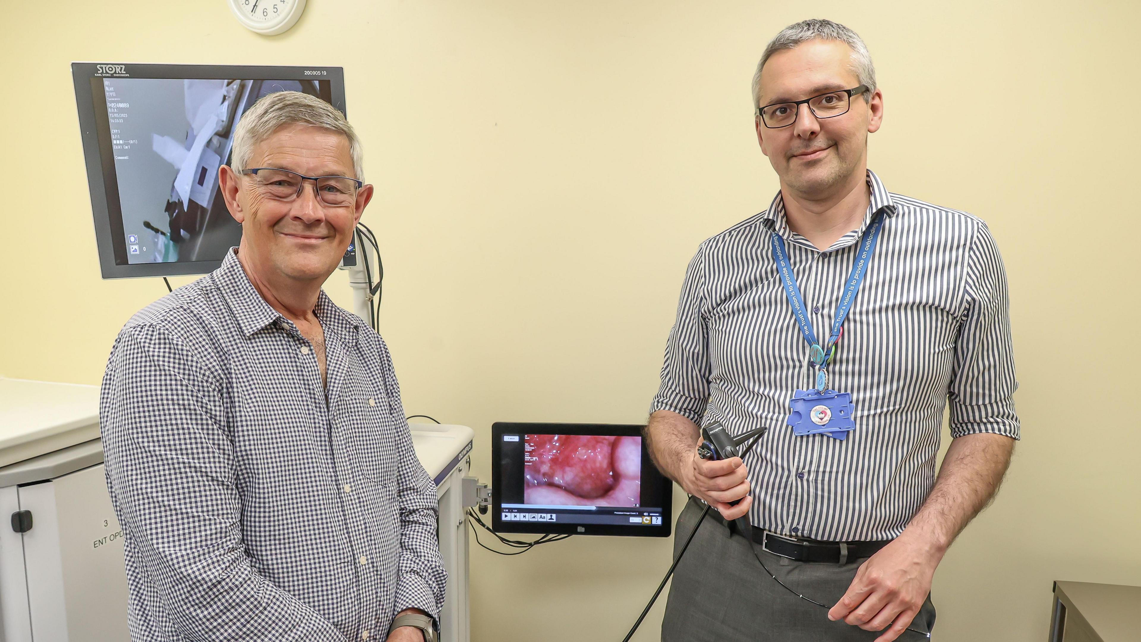 Peter Russel stands next to ENT consultant Mr Kostas Marinakis. Peter is wearing a white and blue striped shirt and has grey hair and glasses. Kostas is also wearing a white and blue striped shirt and has grey hair and glasses. Kostas is holding a scoping device in his hand and in the middle of the two men is a screen which appears to show the inside of someone's throat.