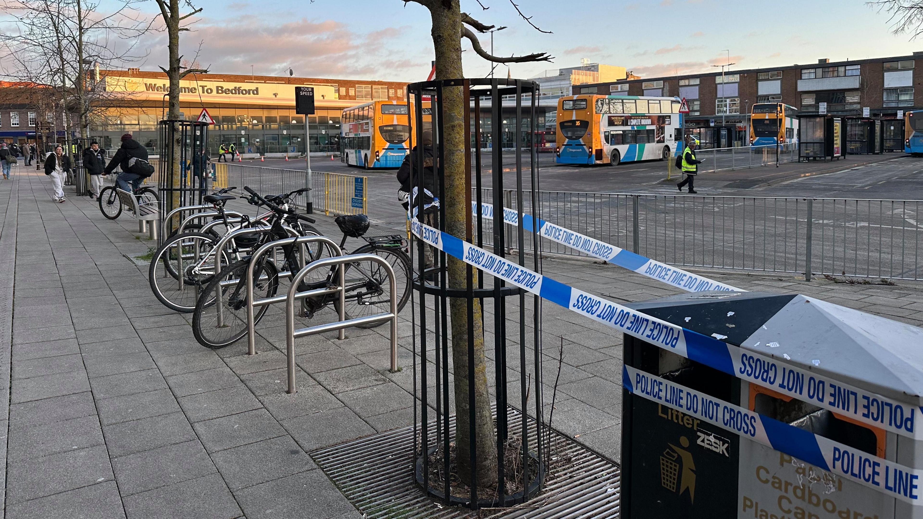 Police tape that says "Police line do not cross" tied round a tree and a bin near Bedford bus station. There is a bike rack next to the cordoned-off tree. Buses are parked outside the bus station.